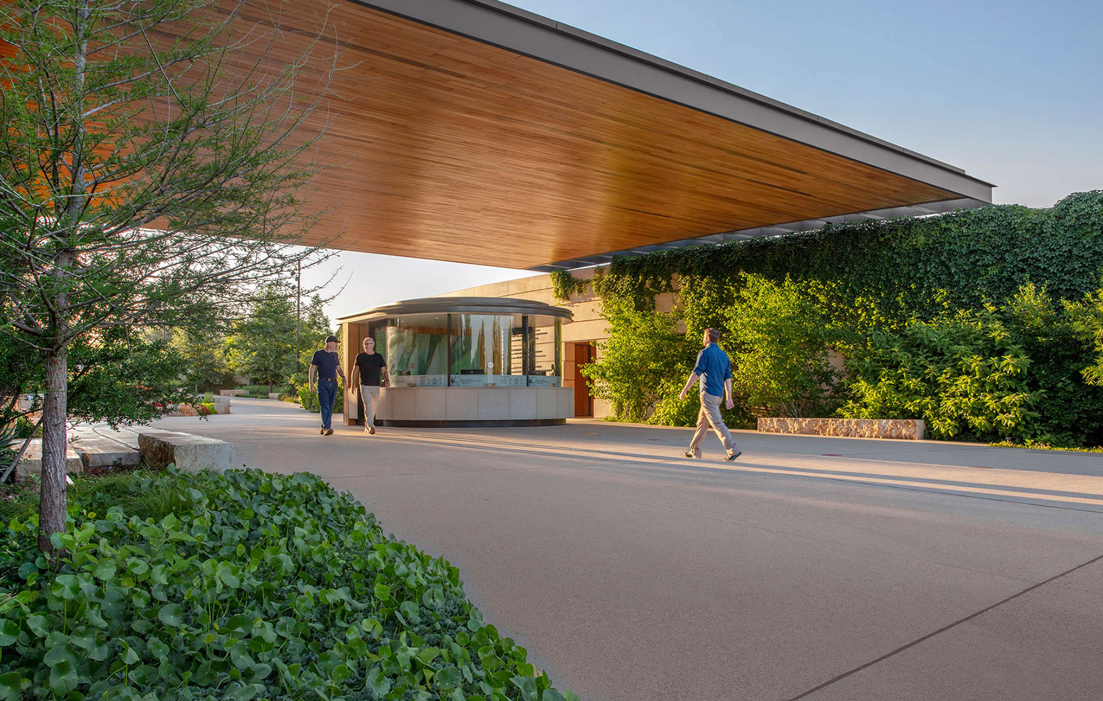 A modern building with an overhanging roof and green ivy walls. People are walking on the pathway in front, with landscaping and trees on either side.