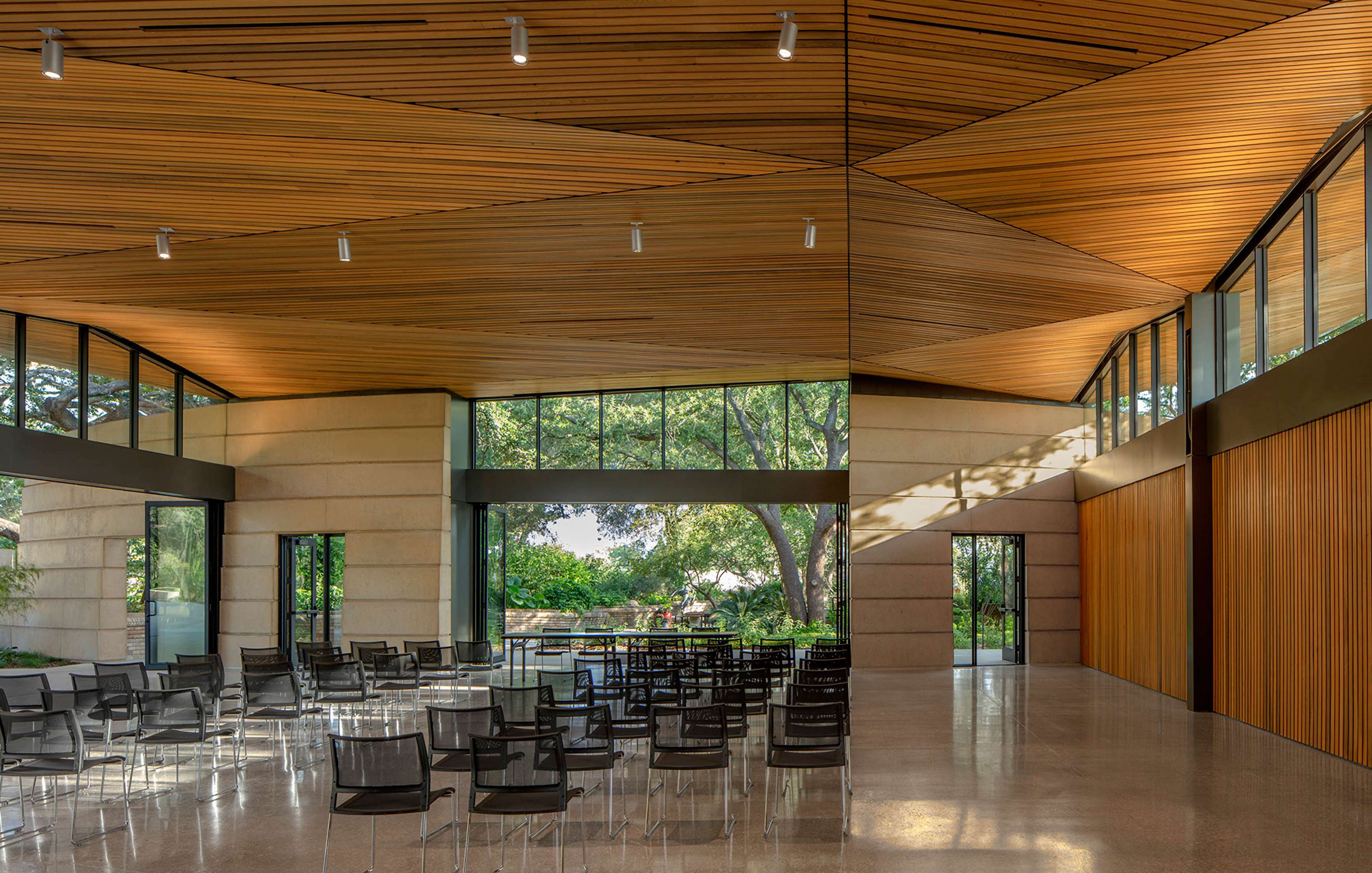 A modern room with a wooden ceiling and large windows, featuring rows of empty black chairs facing forward. Natural light illuminates the space through exterior greenery.