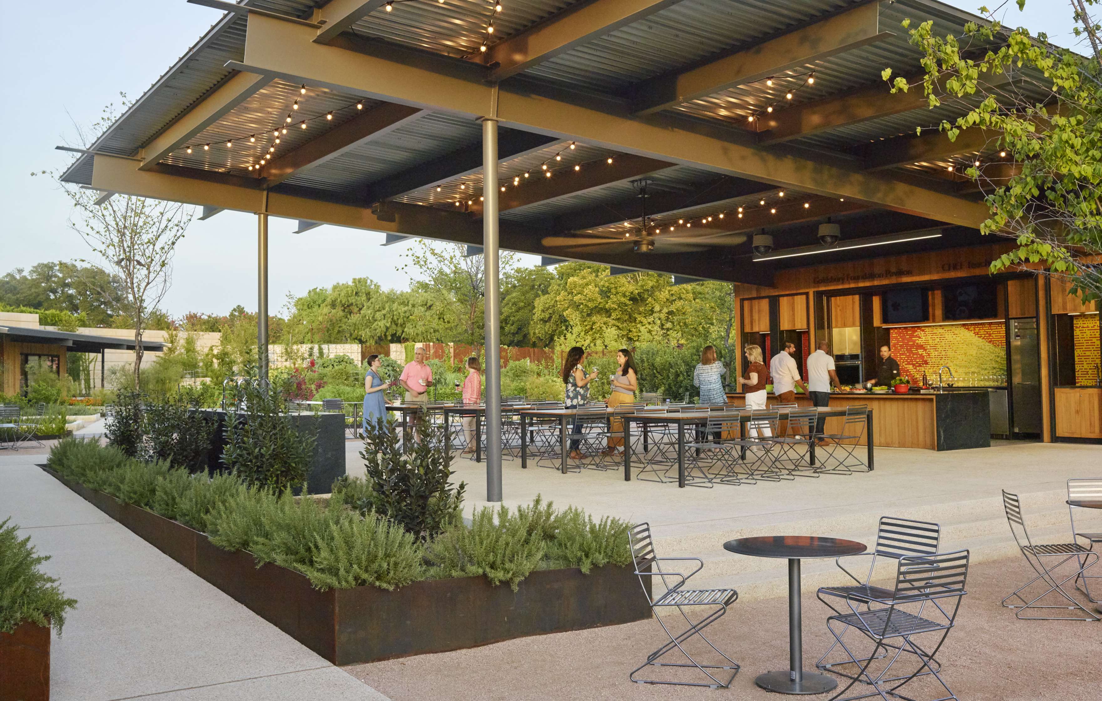 Outdoor bar with string lights, several people standing or sitting around high tables under a covered patio. Greenery and empty tables with chairs in foreground.