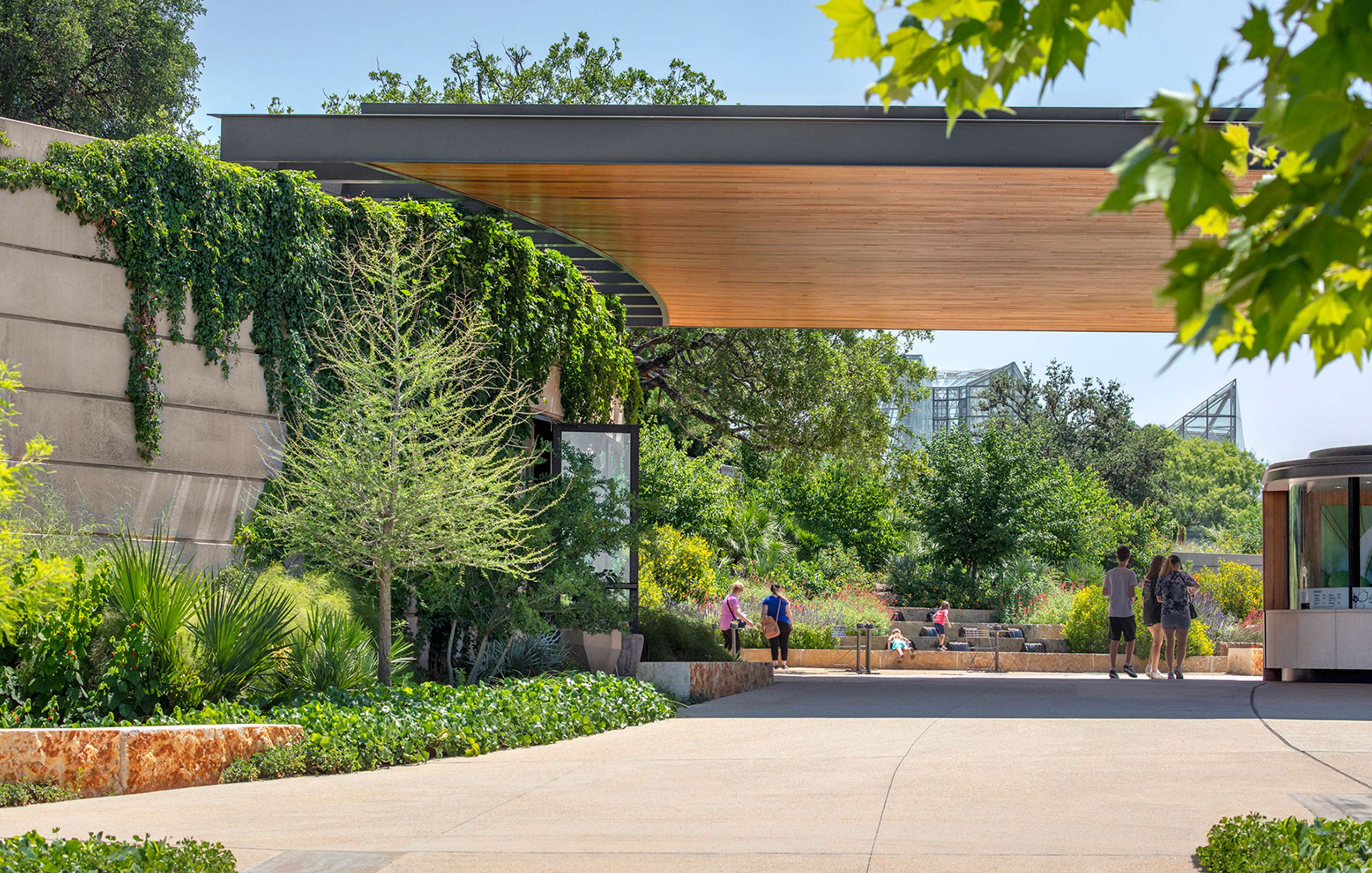 An outdoor garden area with a modern wooden canopy, lush greenery, and visitors walking and resting on benches.