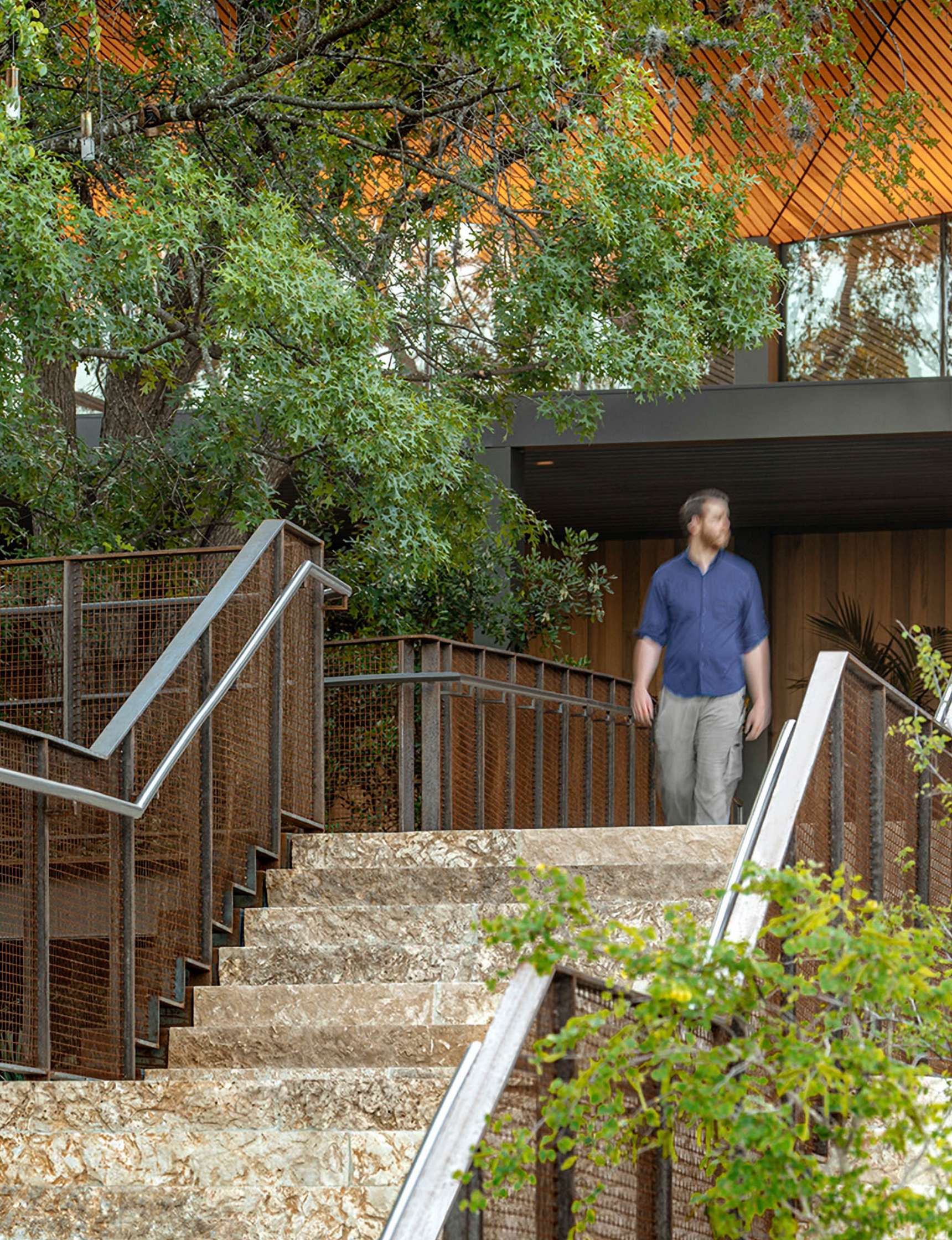 A person walks on a stone staircase bordered by metal railings surrounded by greenery and trees. A building with a wooden exterior and large windows is partially visible in the background.