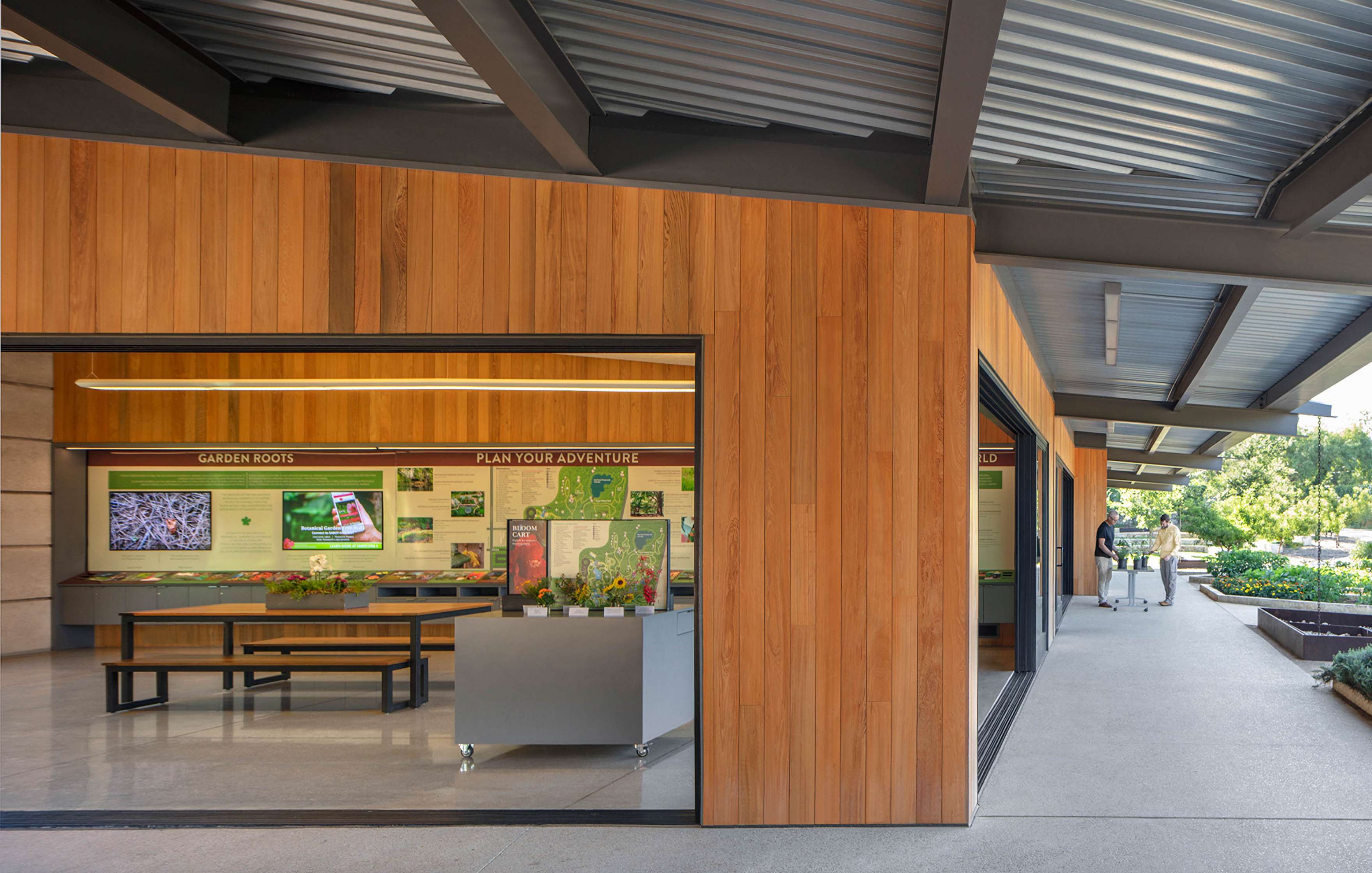 A modern visitor center with wooden panel walls features informational displays about garden roots and adventure planning. Two people walk outside on a paved path. A green landscape is visible in the background.