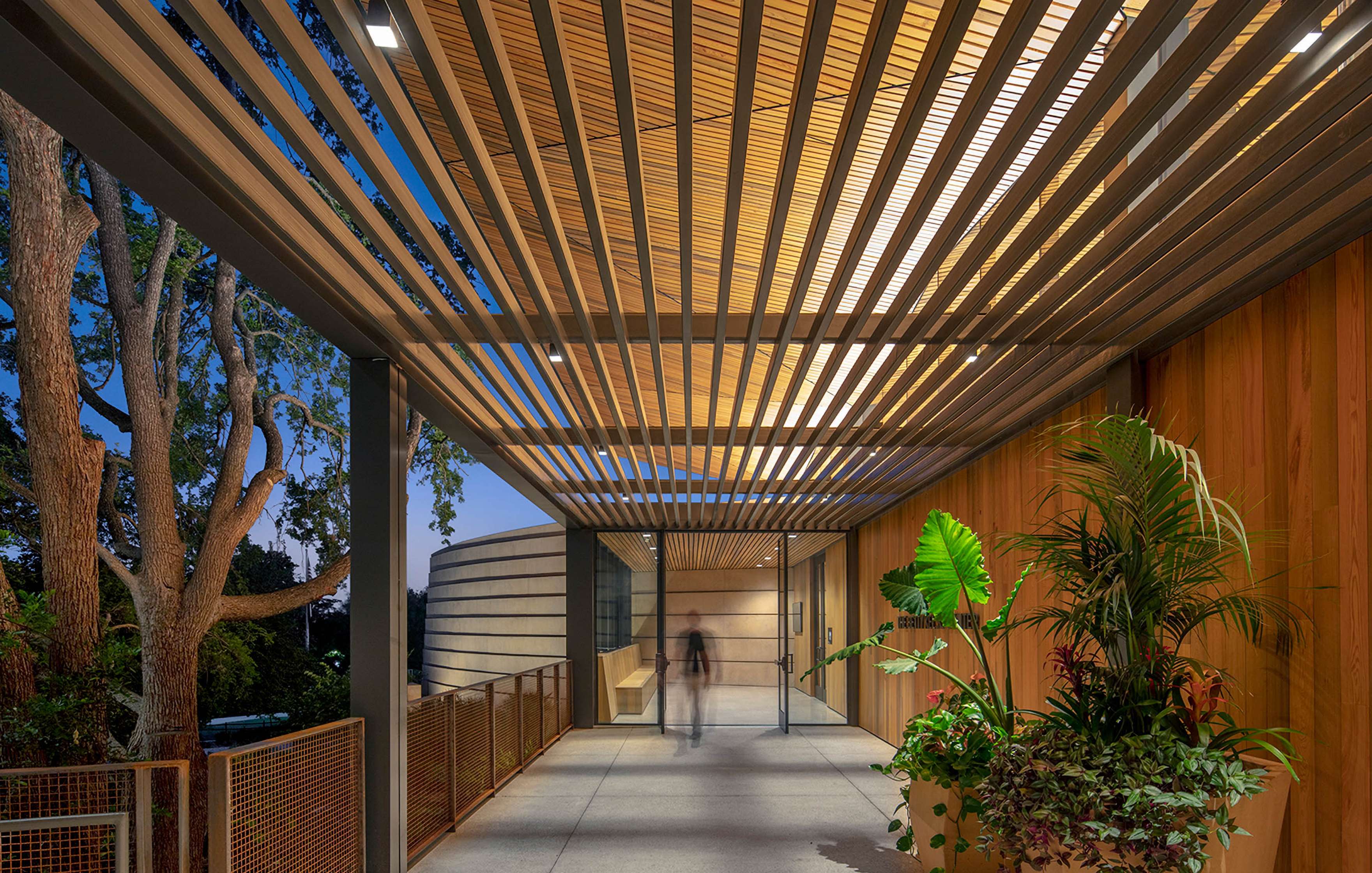 A modern architectural structure with wooden slats, a potted plant, and glass doors. A blurred figure walks through the illuminated, open-air corridor at dusk.