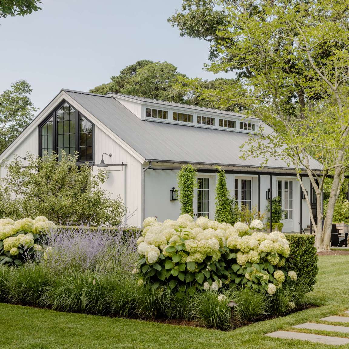 A small, modern house with a gable roof is surrounded by well-manicured landscaping and blooming hydrangeas, situated in a lush green yard with trees in the background.