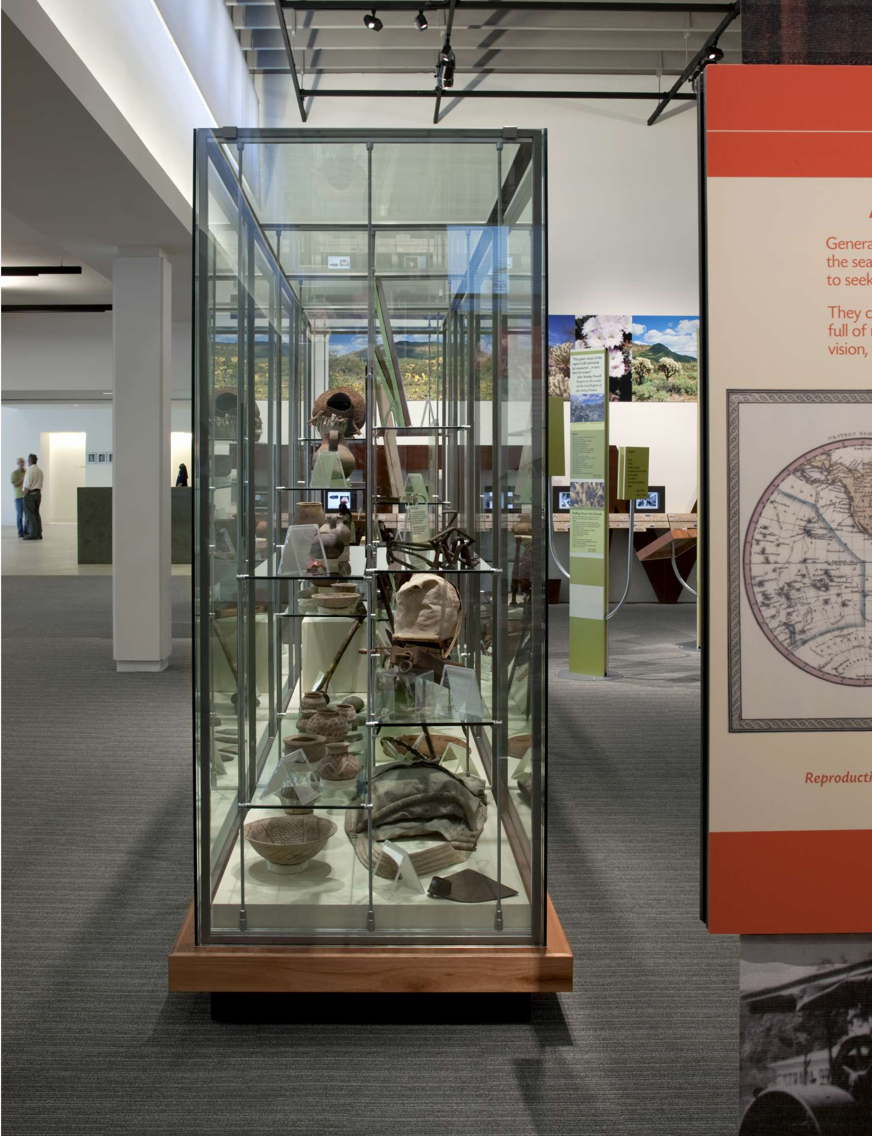 A glass display case contains various artifacts such as pottery and tools in a museum setting. There are information plaques and a world map visible in the background.