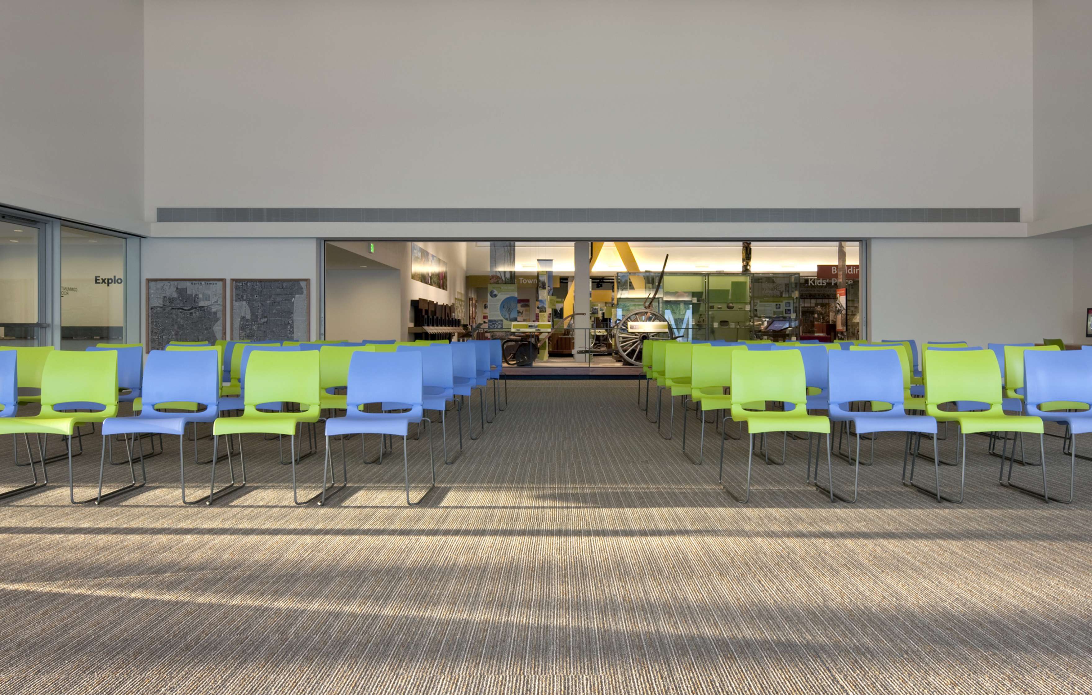 A large room with rows of empty green and blue chairs facing a windowed wall. There is a carpeted floor and a well-lit open area beyond the glass.