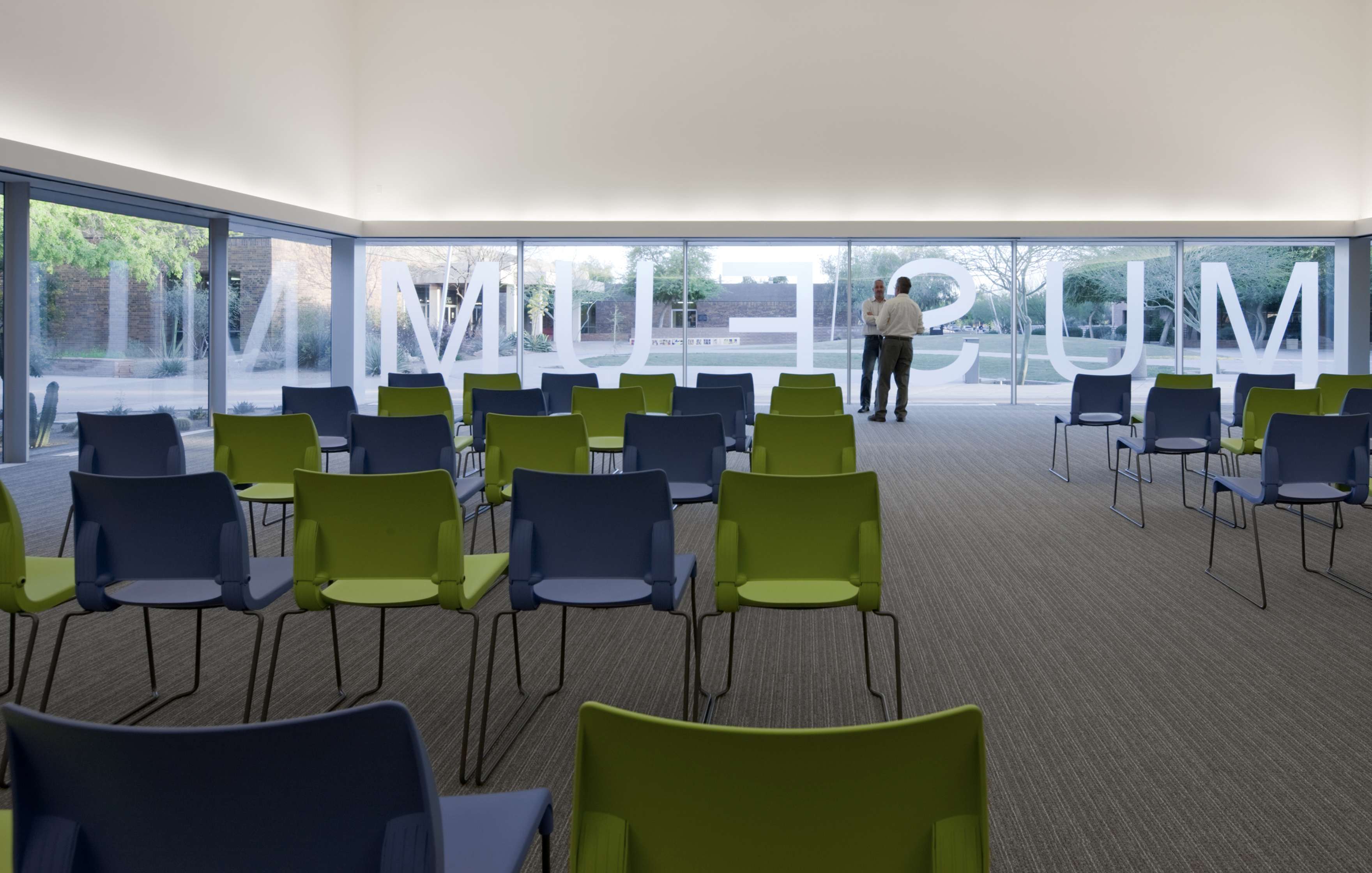 An empty room with rows of green and blue chairs faces a large glass window with the word “MUSEUM” in white letters; two people are standing outside the window.