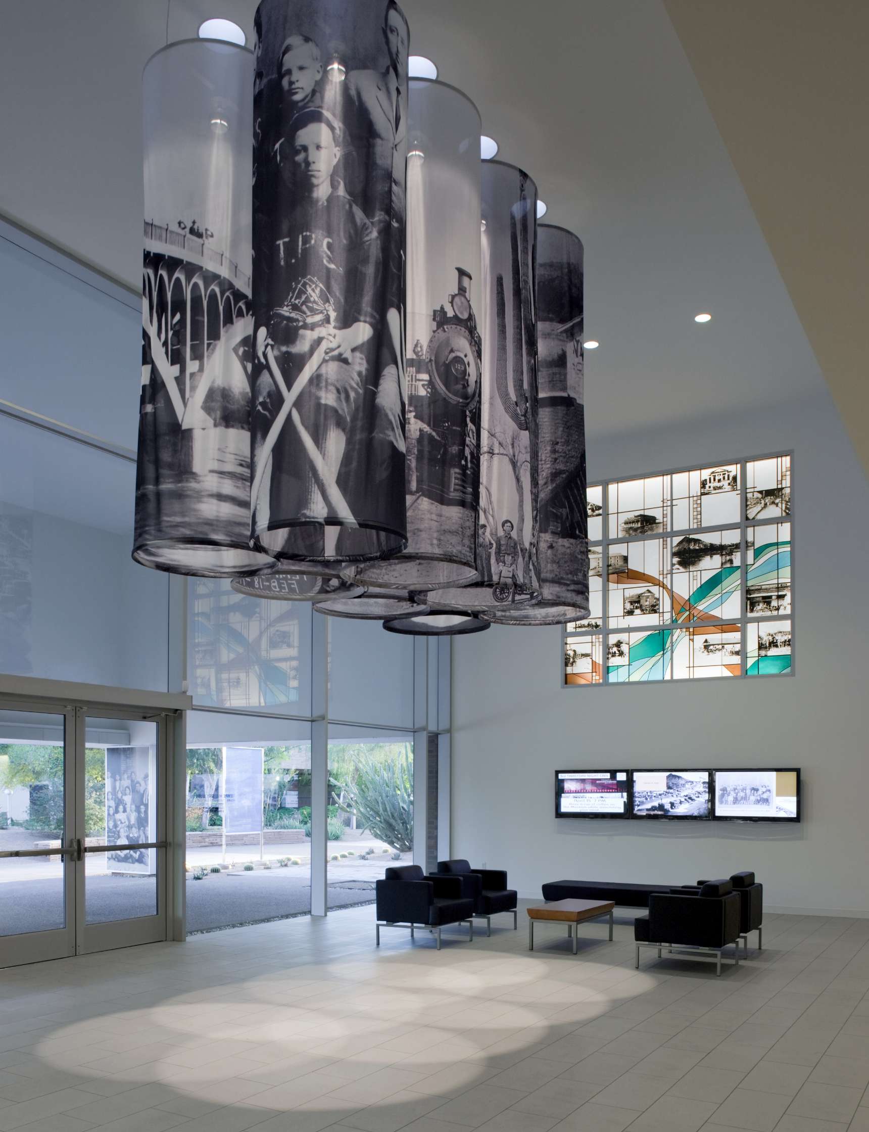 A modern lobby features large cylindrical hanging banners with historical photos, a seating area with black furniture, and multiple display screens on the wall.