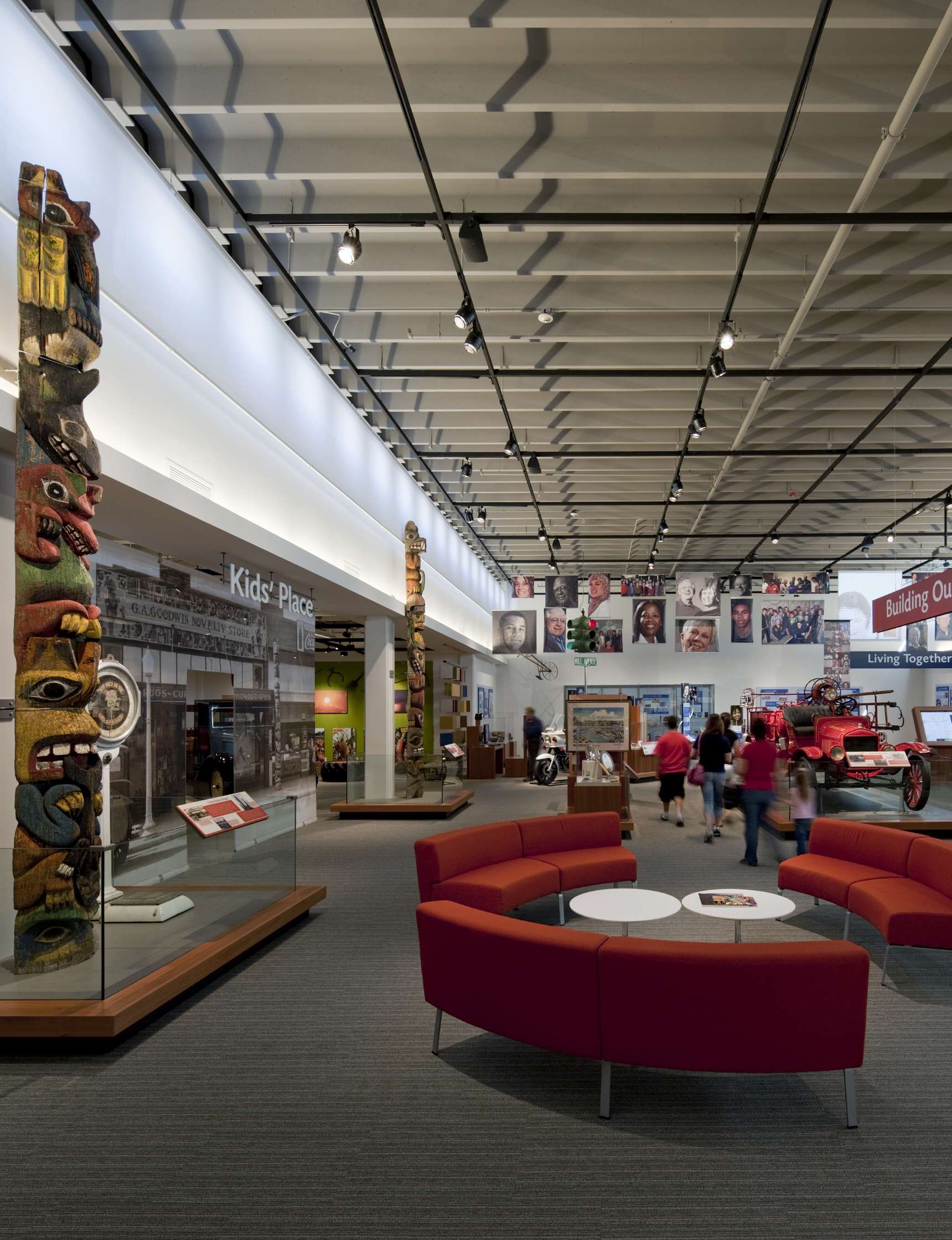 A museum interior featuring exhibits with totem poles, a red circular seating area, and visitors exploring displays.