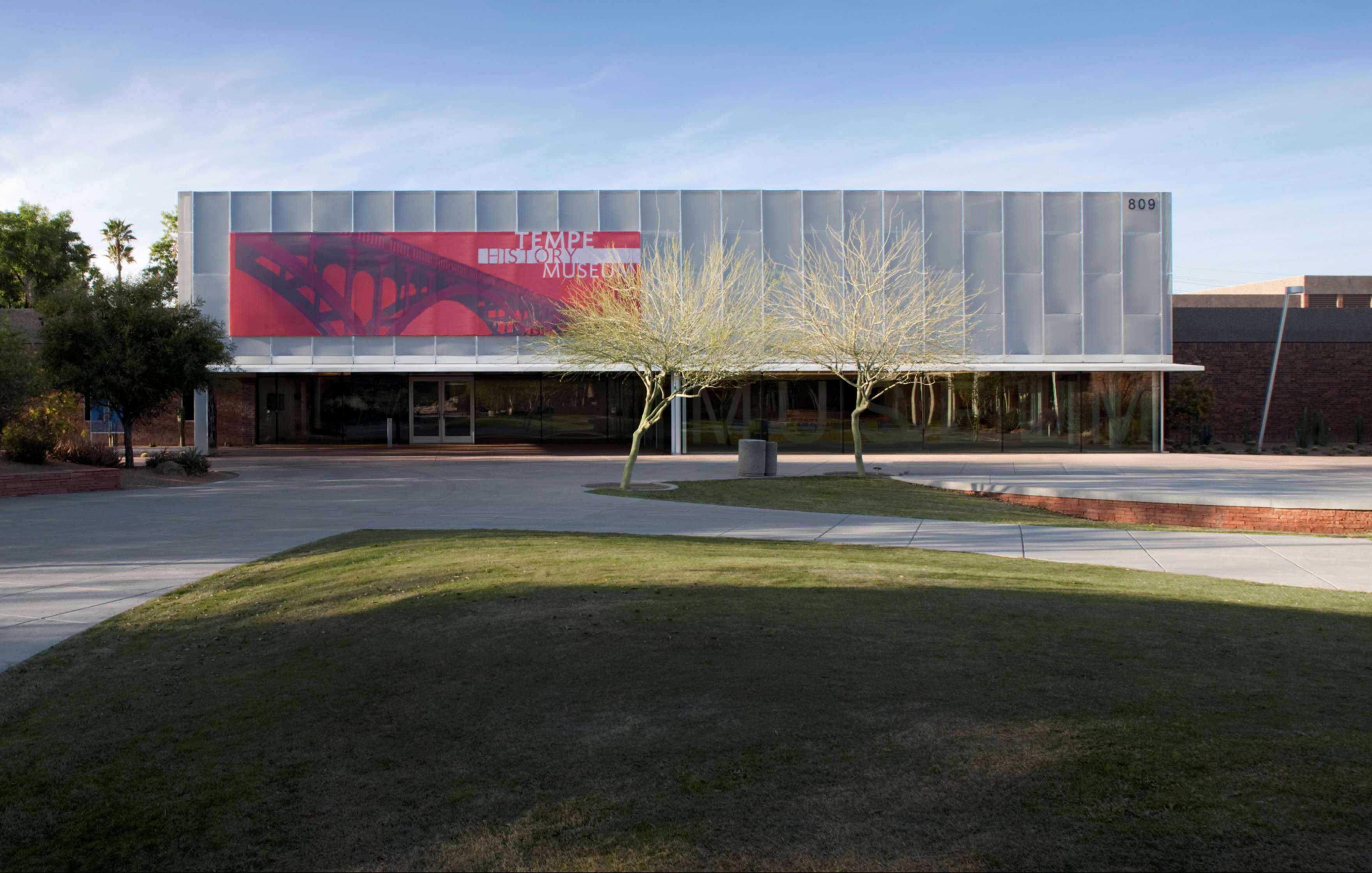 Exterior view of the Tempe History Museum, featuring a modern facade with a red banner on the upper section and a sidewalk leading to the entrance. Trees stand in front of the building.