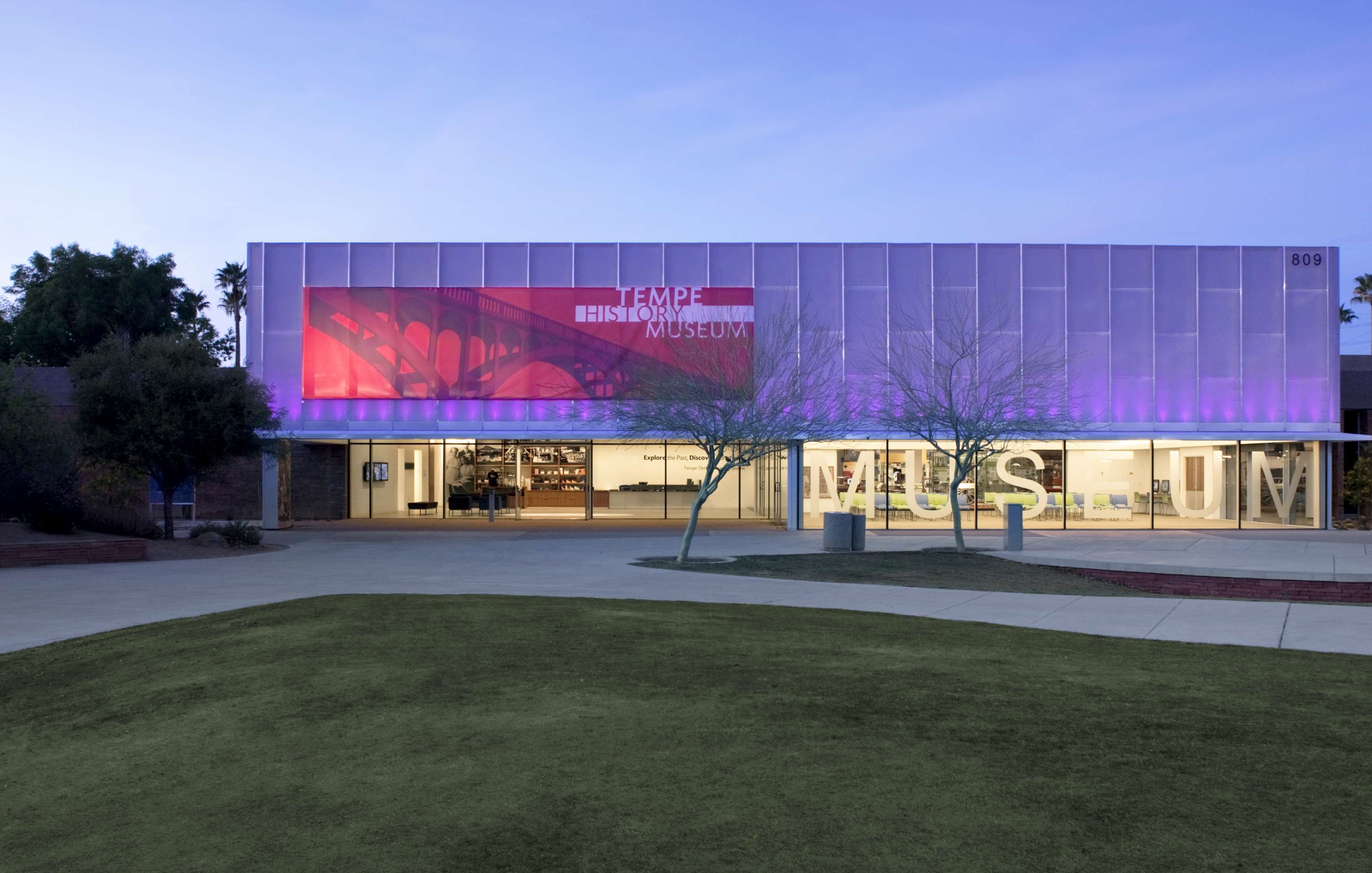 A modern building with a bold red sign reading “Tempe Historical Museum” on its facade, displaying well-lit interior and exterior at dusk. The entrance is flanked by landscaping and a walkway.