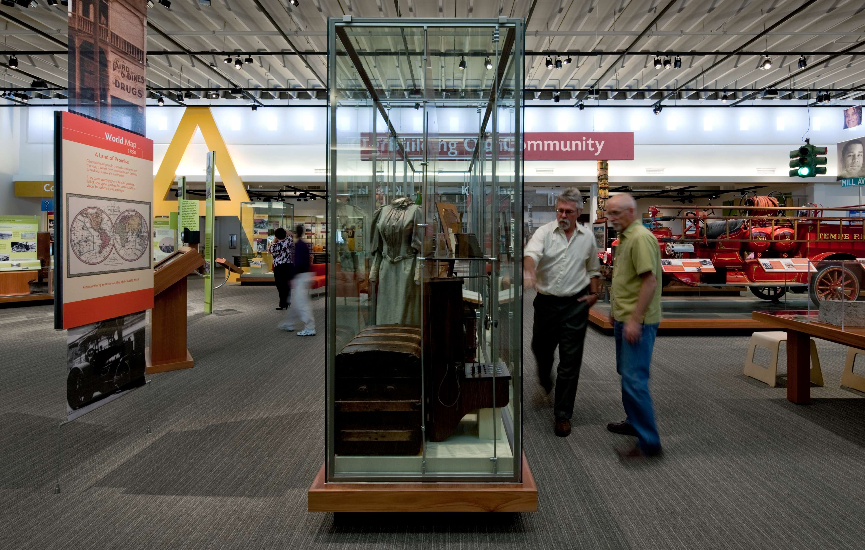 Two people are viewing a glass display case with historical artifacts in a museum exhibit. Various other displays and exhibits are visible in the background.