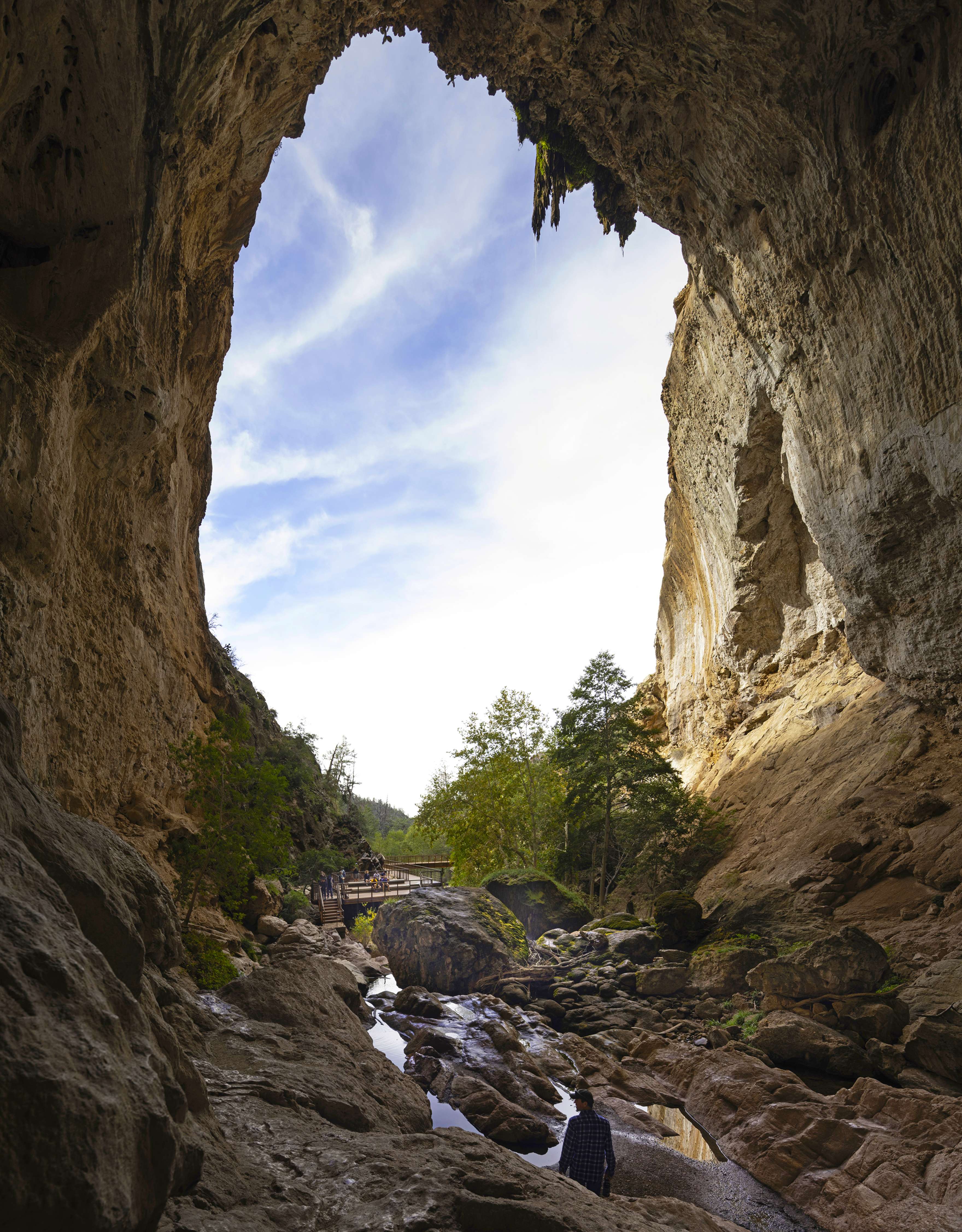 A view from inside a cave looking out towards a rocky landscape with trees and a small footbridge in the distance under a partly cloudy sky.