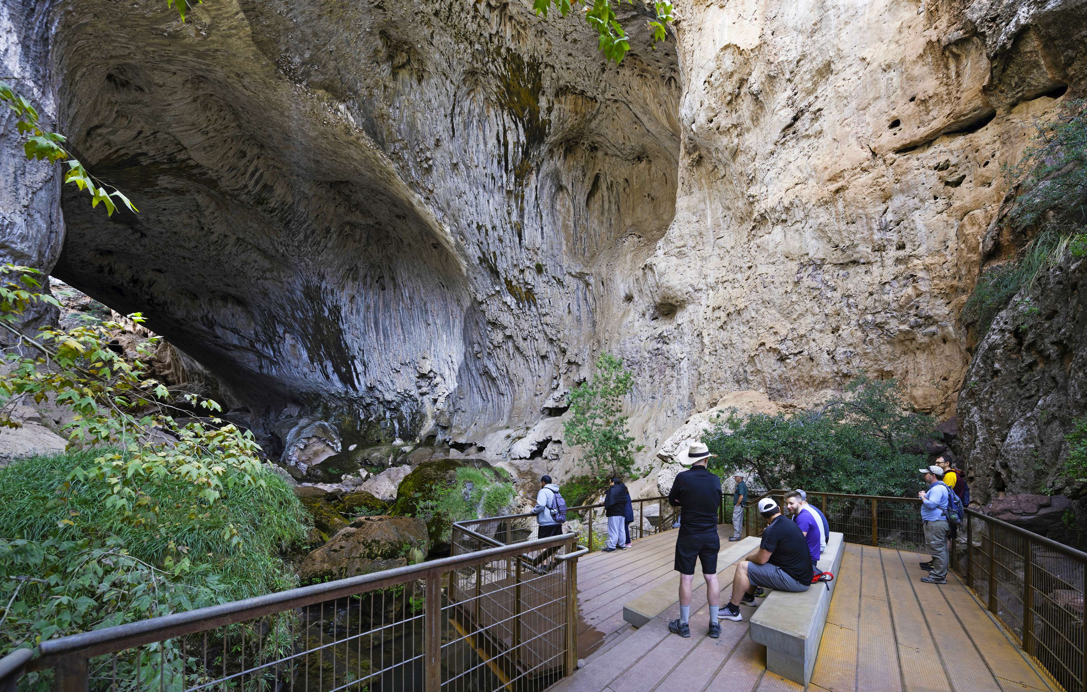 A group of people stand and sit on a wooden deck, observing the large rock formations inside a cave with greenery around.