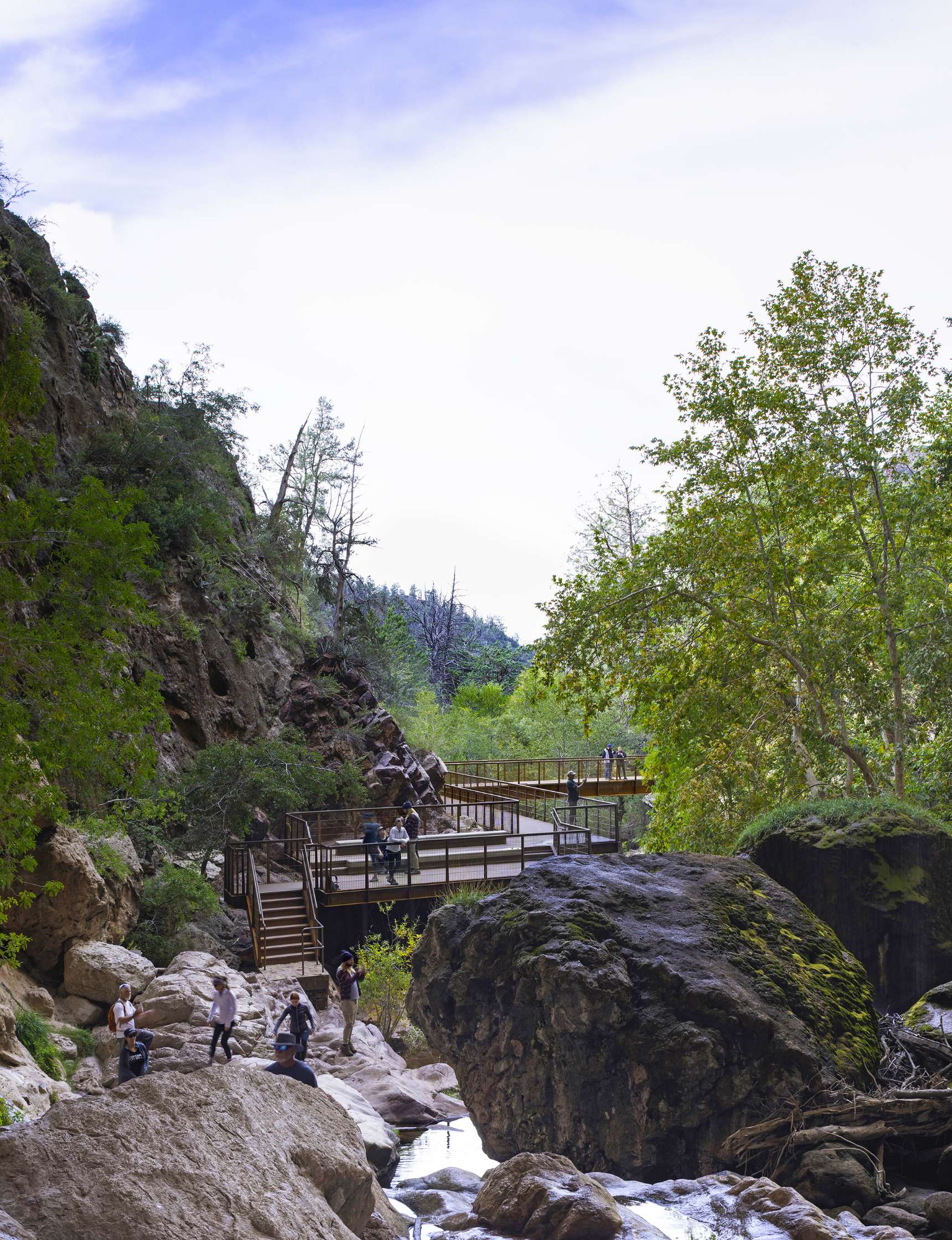 Visitors explore a scenic nature trail with wooden platforms and stairs amidst rocky terrain and lush greenery.