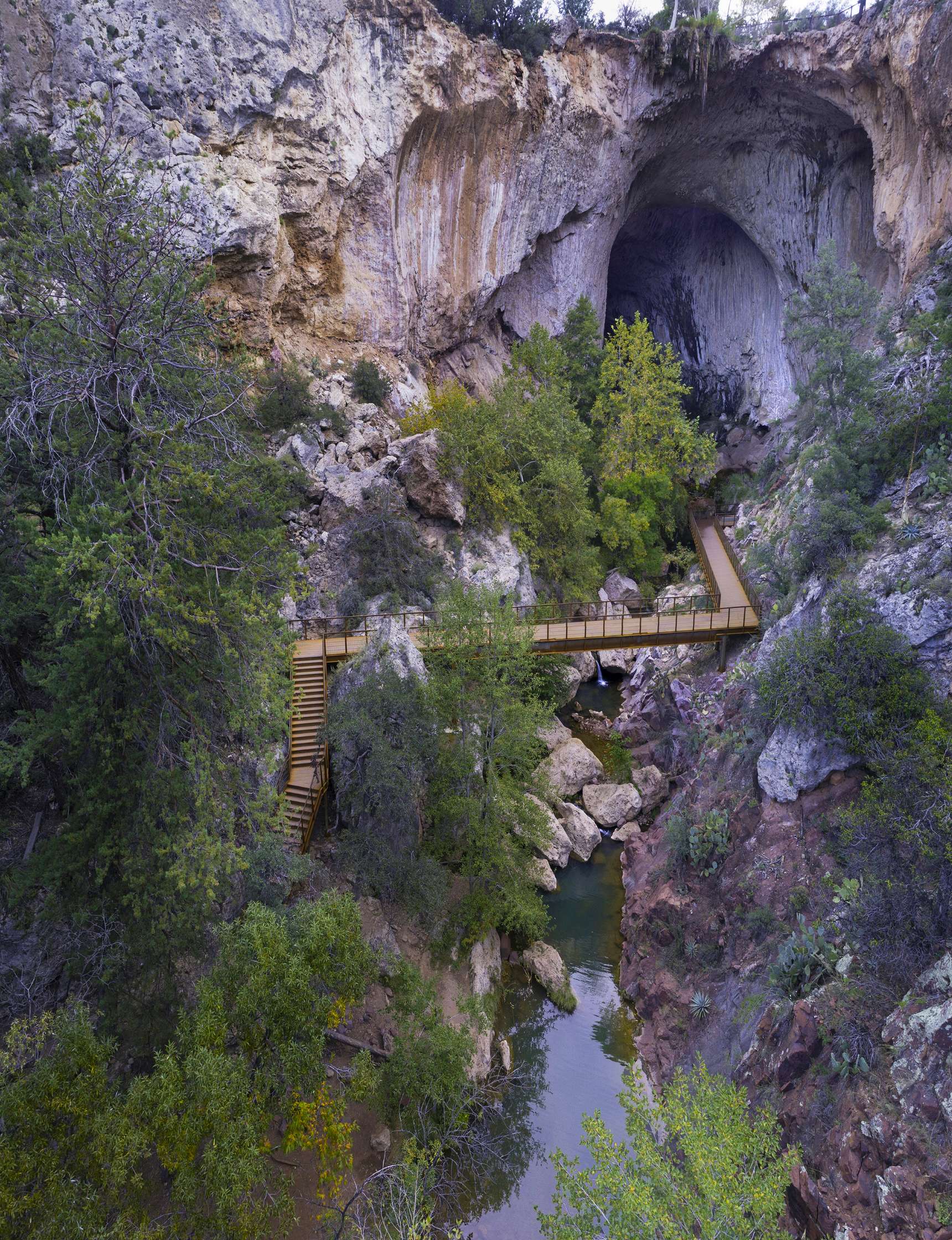 Rock formation with a large cave opening and a wooden walkway bridge crossing over a stream surrounded by dense greenery.