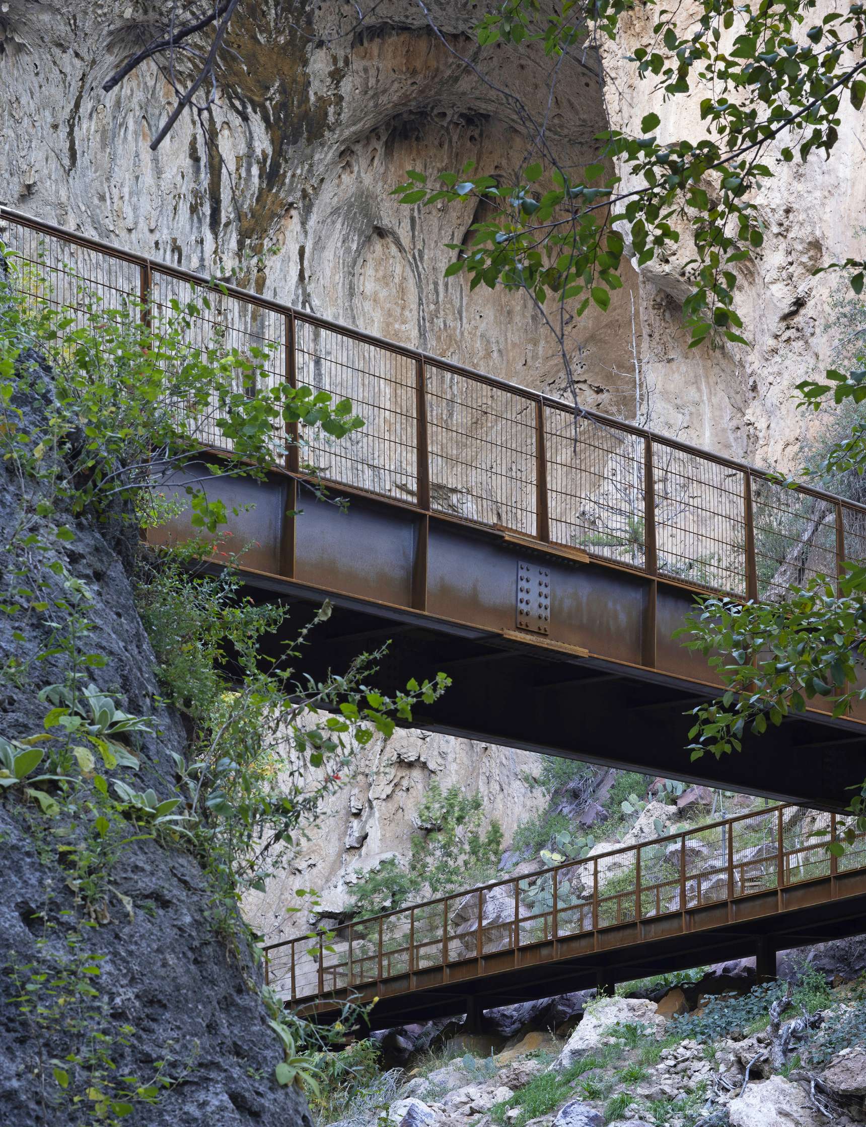 Two rusted metal bridges span between rocky cliffs in a forested area with lush green foliage.