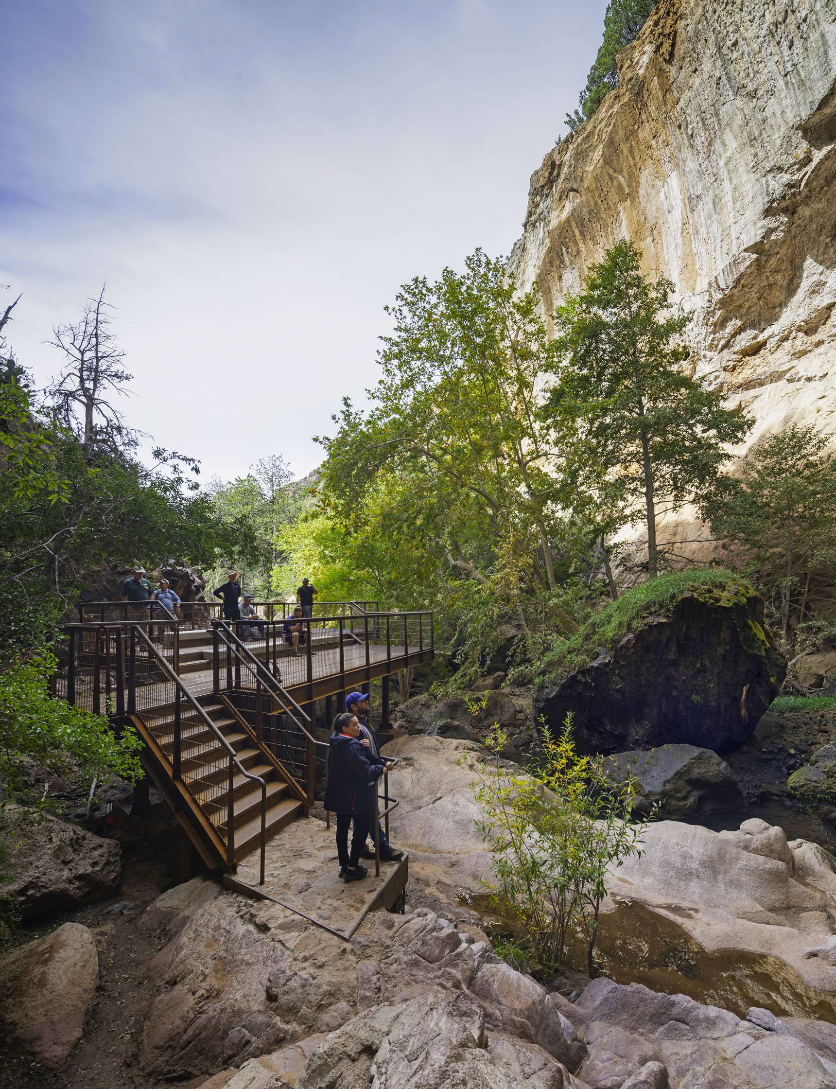 Visitors stand on a viewing platform, looking at the natural landscape and rock formations in a canyon. The scene includes trees, rocks, and a cloudy sky.