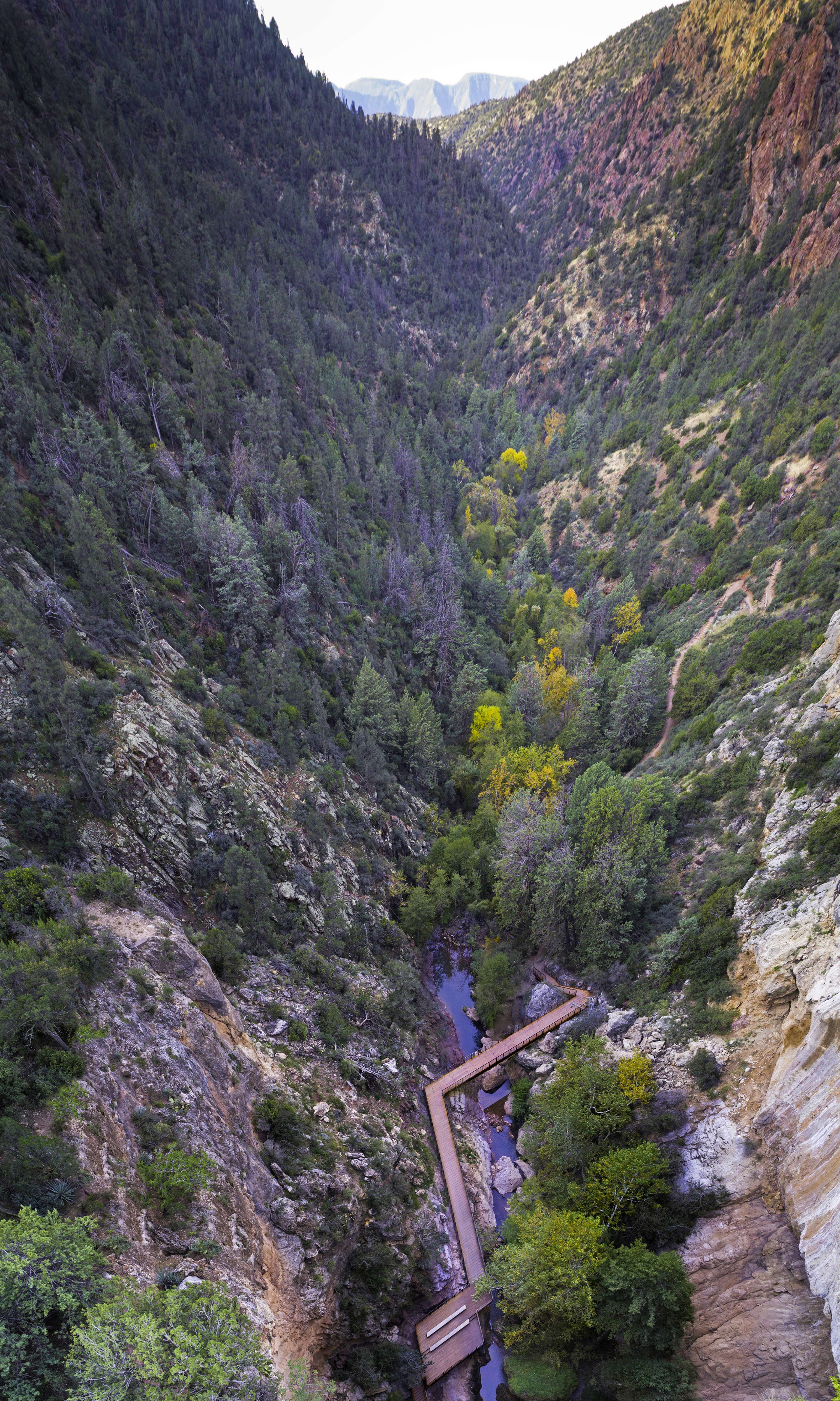 A wooden bridge spans a narrow river within a deep, forested valley surrounded by steep, rocky hills.