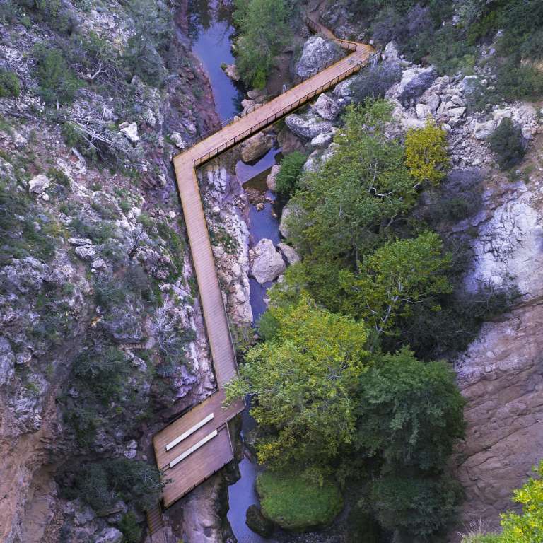 A wooden walkway and bridge traverse over a narrow rocky gorge, surrounded by greenery and streams below.