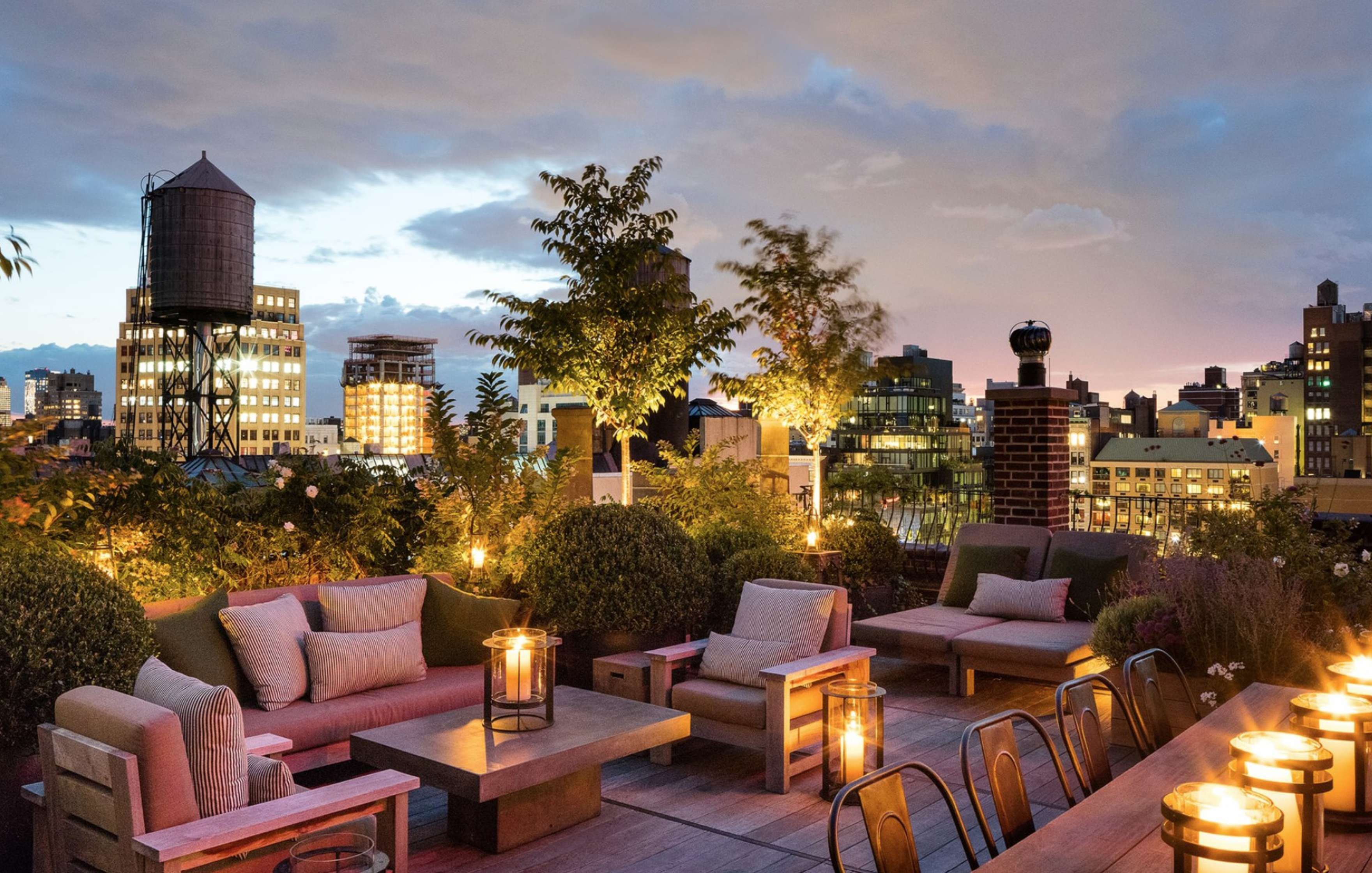 A cozy rooftop terrace at dusk with cushioned seating, lanterns, plants, and a cityscape featuring buildings and a water tower in the background.