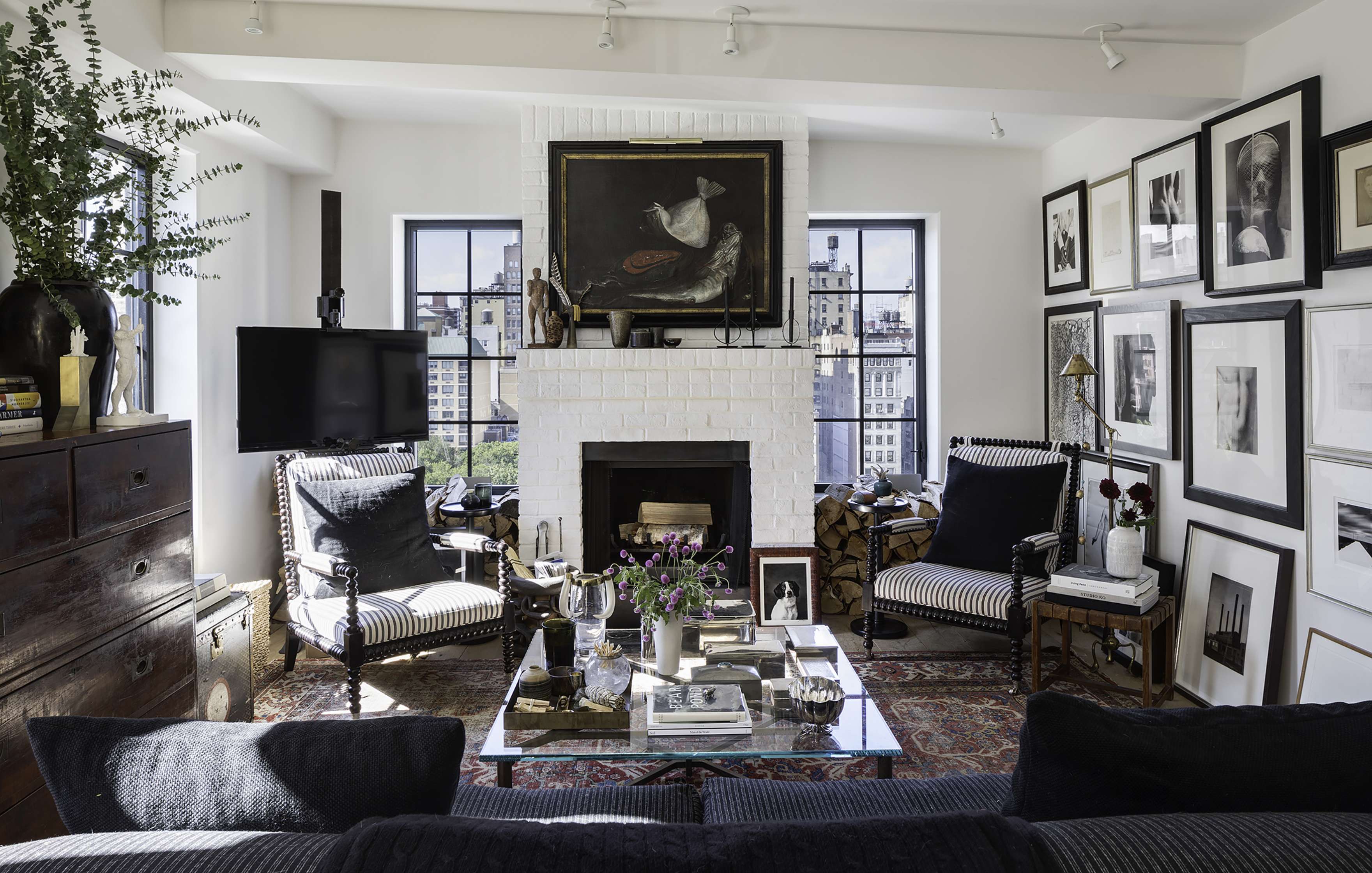 A living room with a white brick fireplace, black framed art on the walls, two striped armchairs, a wall-mounted TV, and a black couch facing a glass coffee table. Large windows offer a city view.