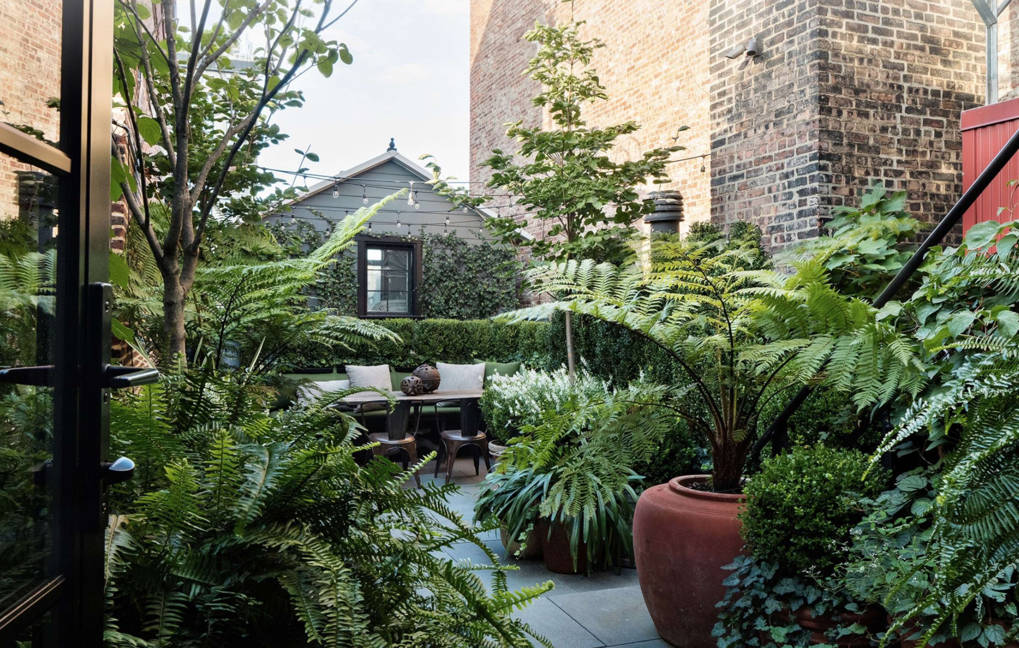 A lush garden with various plants and ferns surrounds a small seating area with a table and chairs, bordered by brick walls and outdoor staircases.