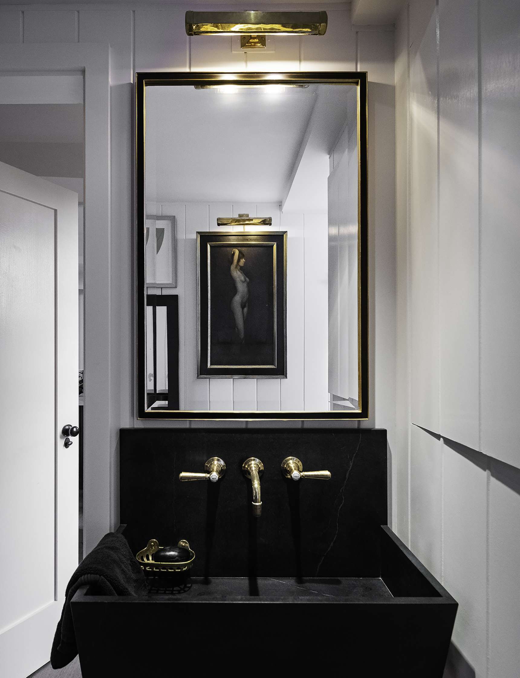 A modern bathroom with a black sink, gold fixtures, a large rectangular mirror, and a framed artwork of a nude figure reflected in the mirror. A neatly folded black towel is placed beside the sink.