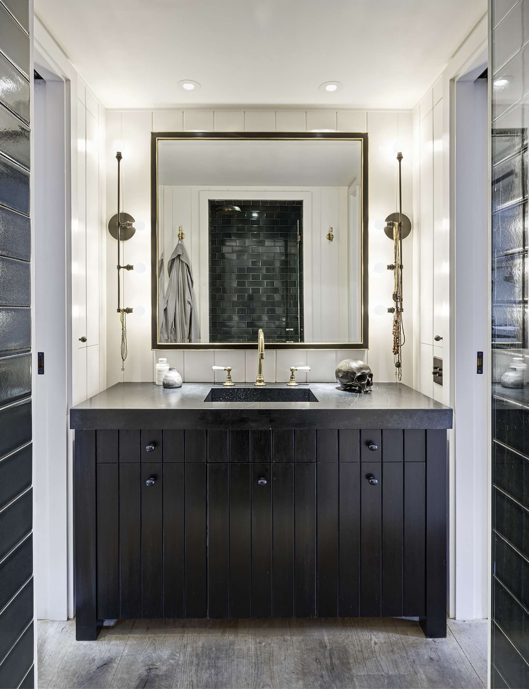 Modern bathroom with black vanity, vessel sink, and brass fixtures. Large framed mirror above the sink, flanked by wall-mounted sconces. Dark tile shower visible in the reflection.