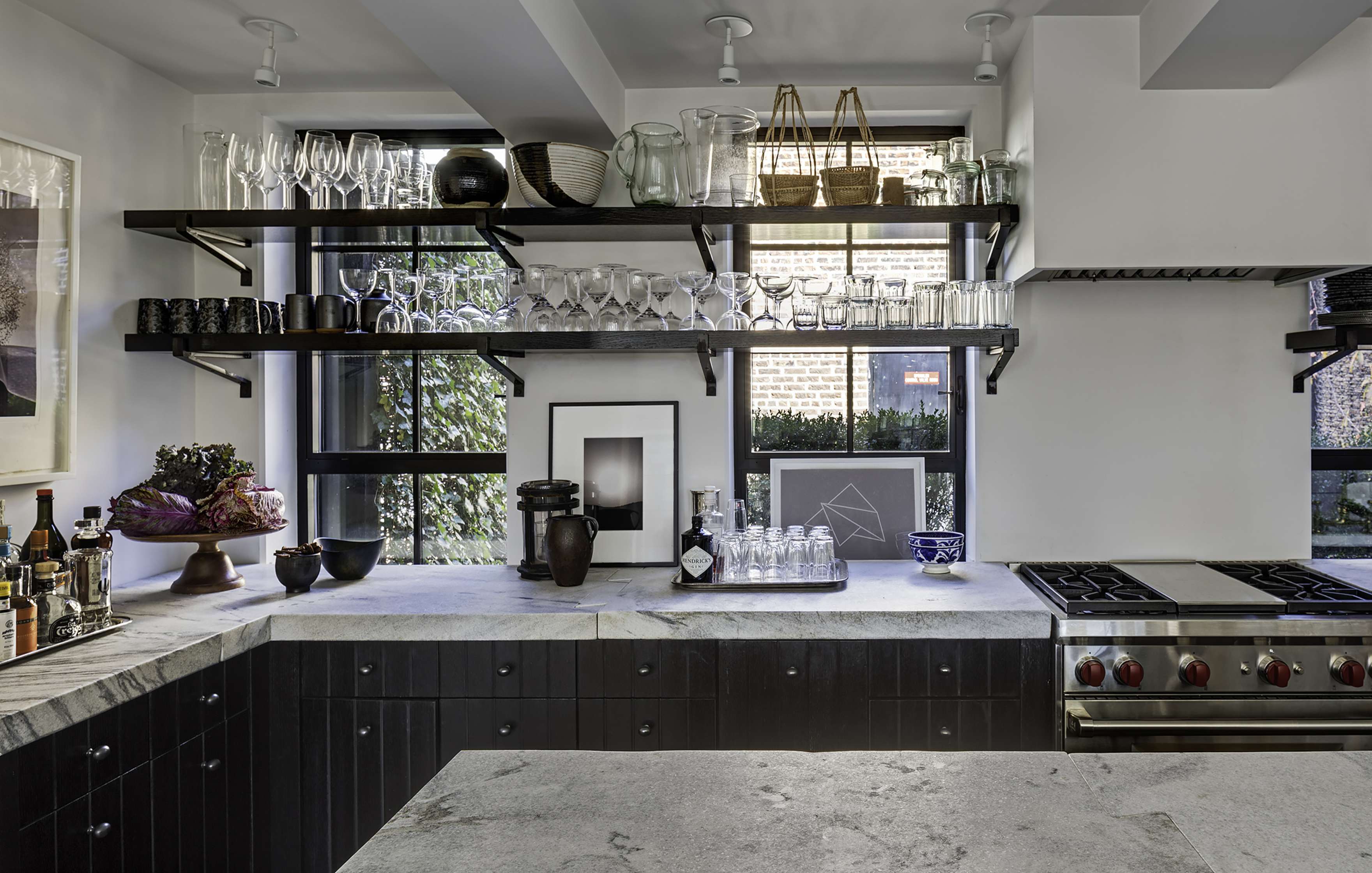 Modern kitchen with dark countertops and cabinets, open shelves holding glassware, and stainless steel appliances. Several decorative items and plants are placed on the counters.