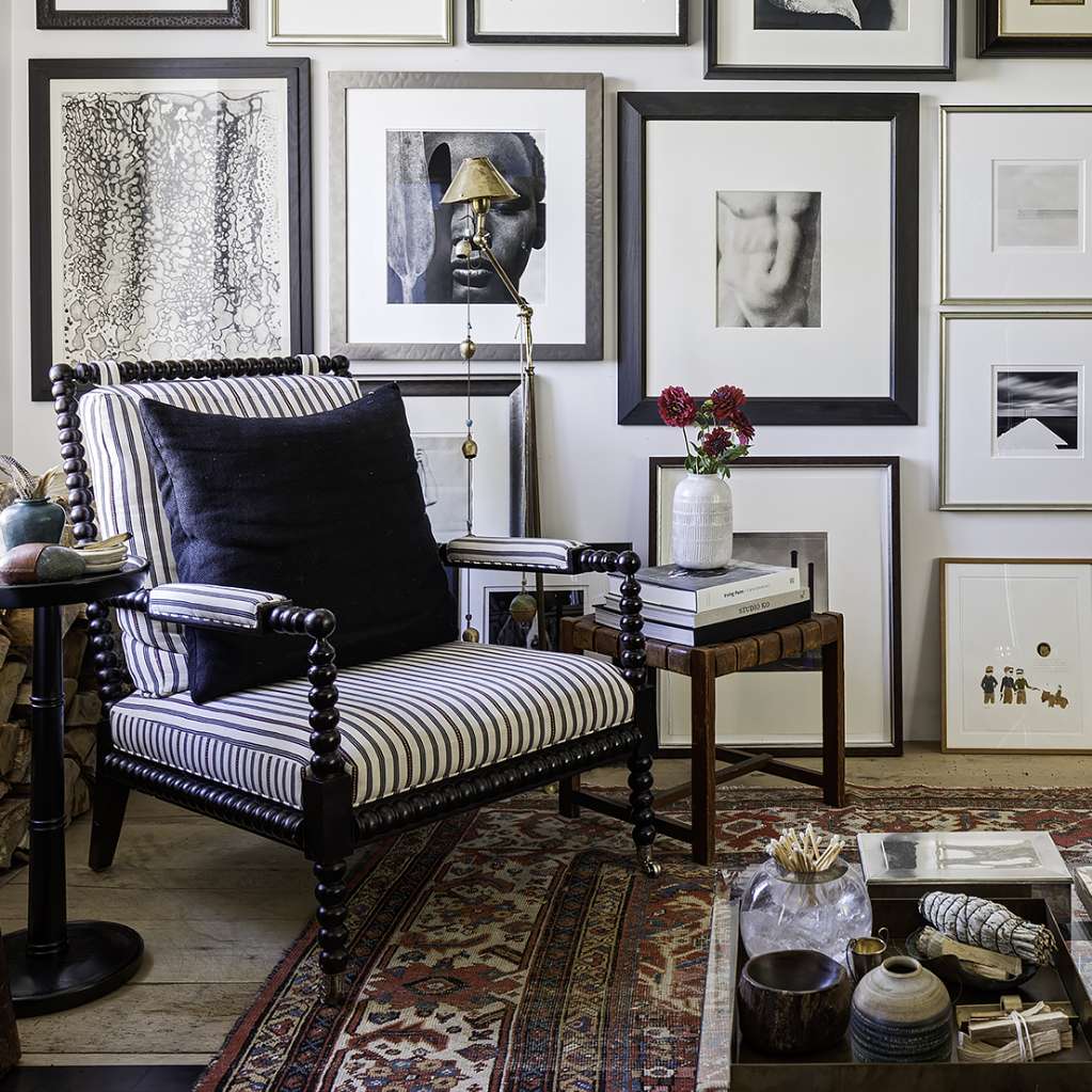 A cozy sitting area features a striped armchair with a black pillow, a side table with decor, and a gallery wall of framed artwork and photographs. A patterned rug and various books are also present.