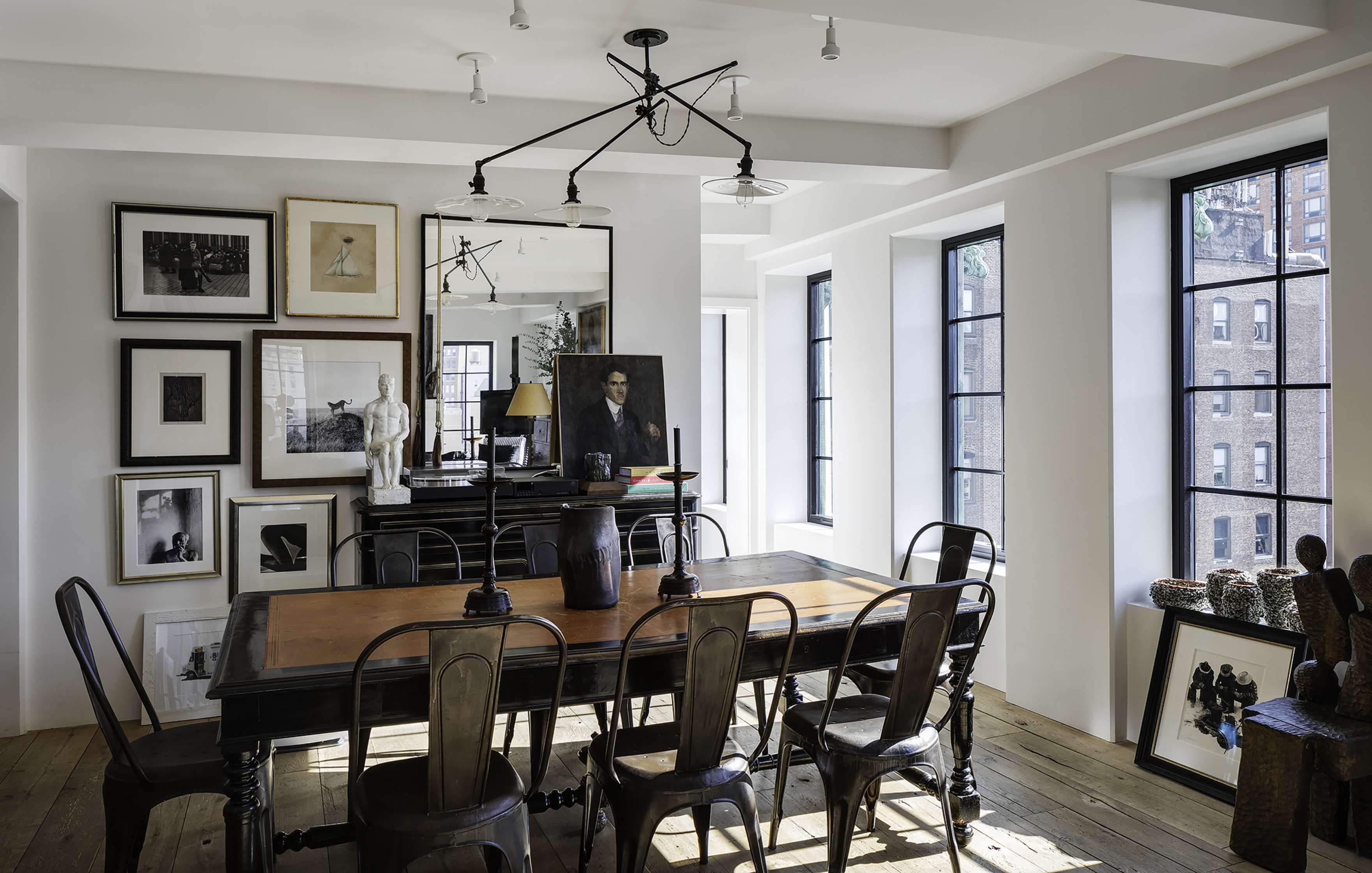 A stylish dining room featuring a wooden table, metal chairs, various framed artworks on the walls, and large windows letting in natural light.