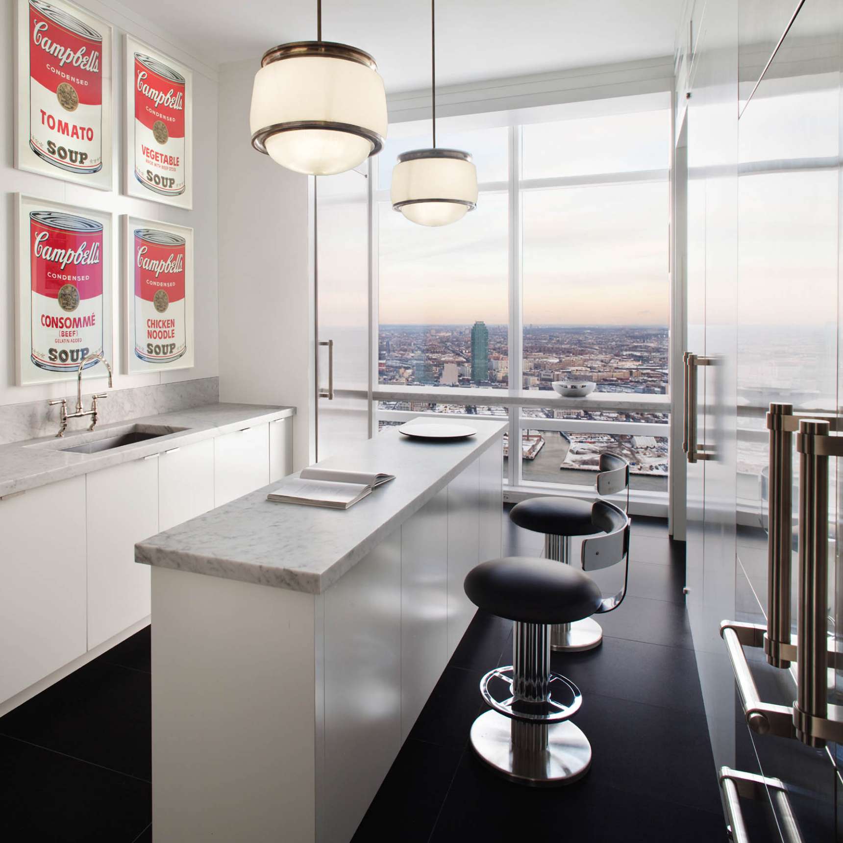 Modern kitchen with a large window offering a city view, featuring a white island with two black barstools, light fixtures, and framed Campbell's soup art on the wall.