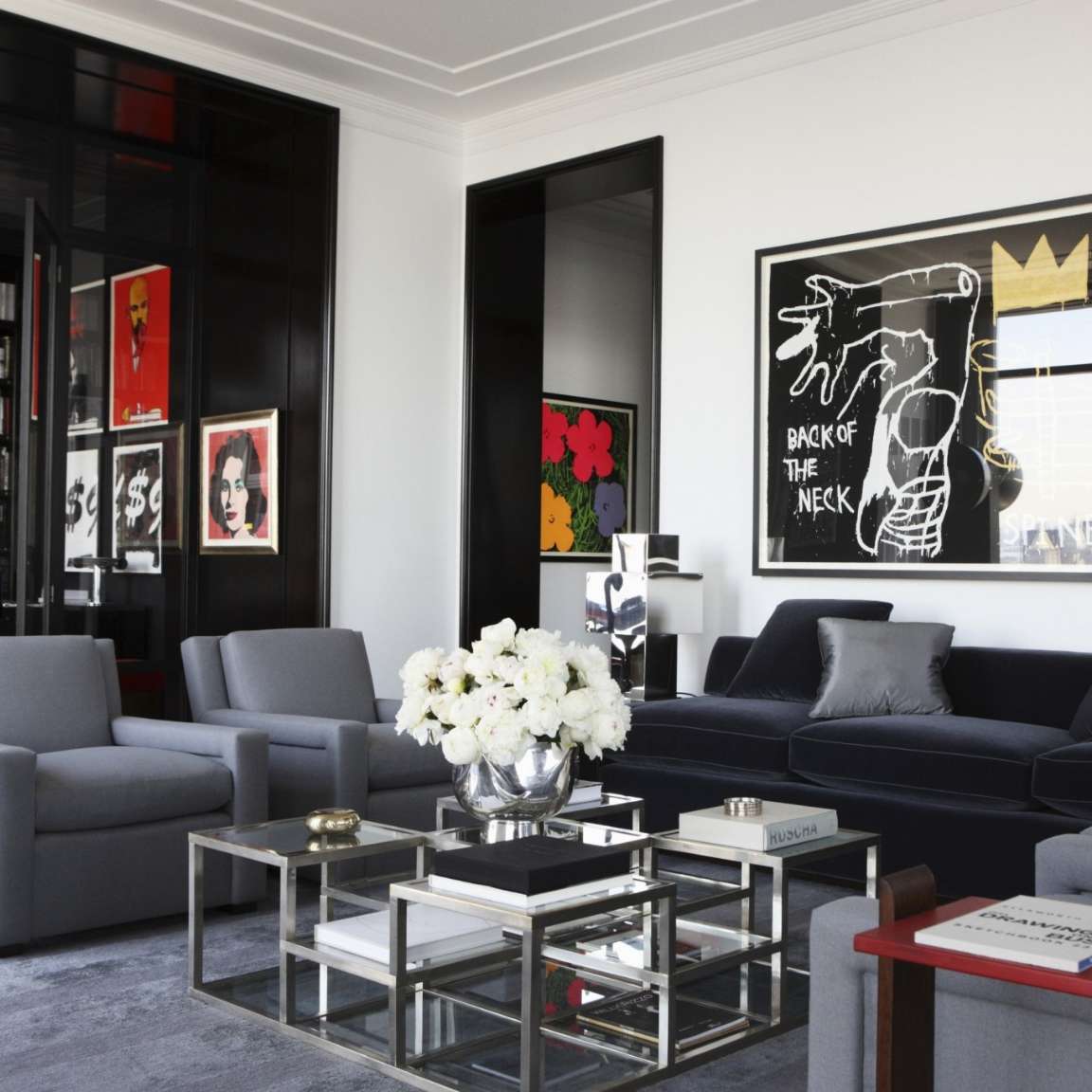 A modern living room with a black sofa, gray armchairs, glass coffee table, and wall art. A vase of white flowers is on the table, and a black and white artwork is hung on the wall behind the sofa.