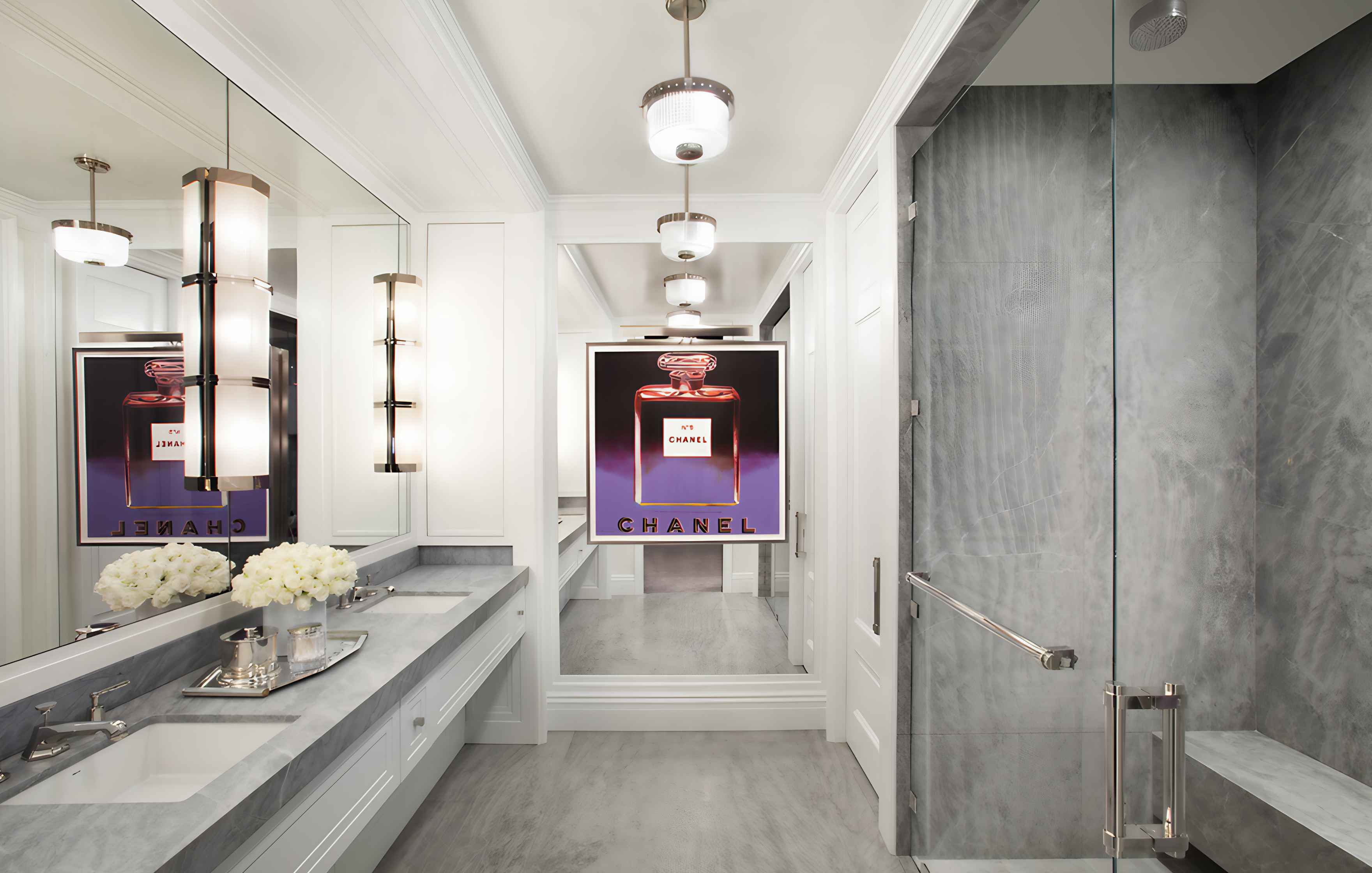 Luxurious bathroom with marble countertop, chrome fixtures, white cabinets, large mirror, and modern lighting. An oversized Chanel No. 5 poster is reflected in the mirror.