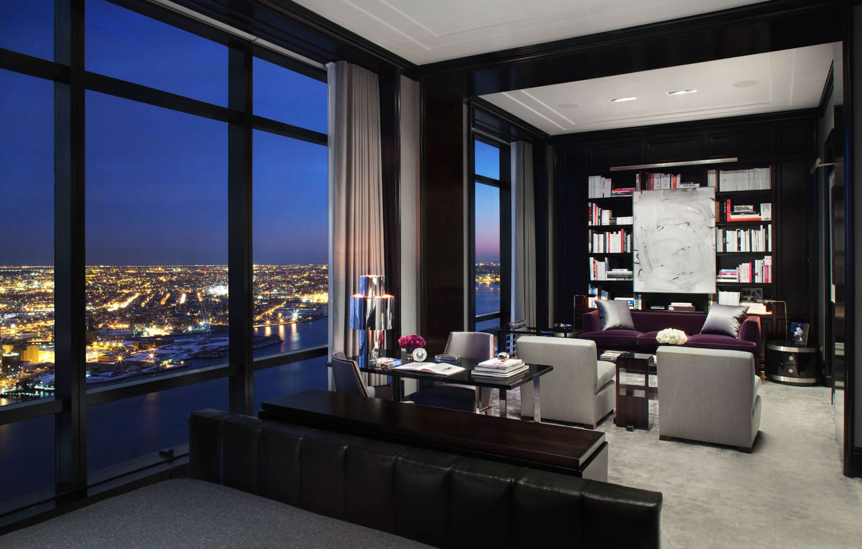 Luxurious modern living room with floor-to-ceiling windows offering a night view of a city's lights. Room includes a sofa, armchairs, bookshelves, and contemporary decor.