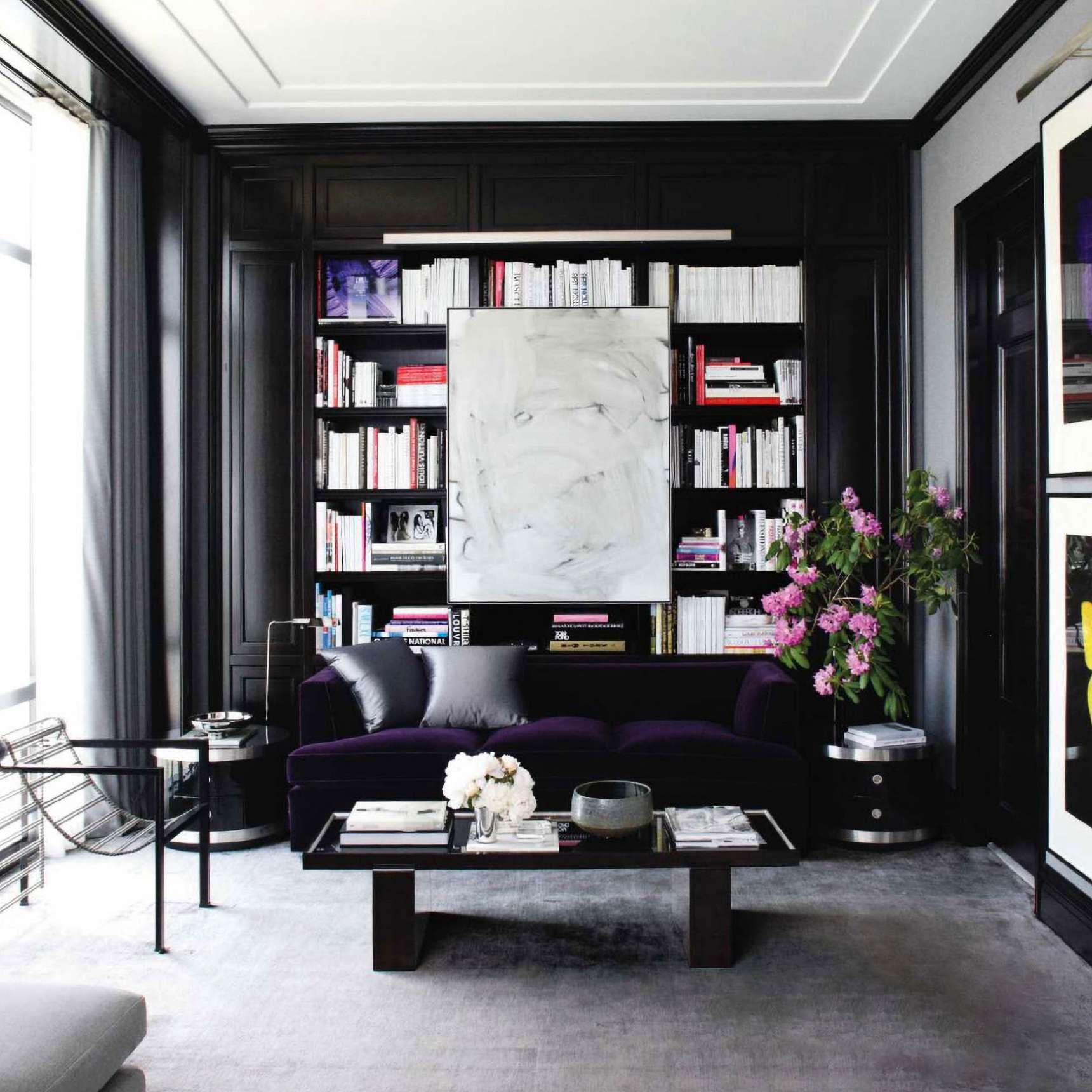 A modern living room with dark wood paneling, a black sofa, a glass coffee table, shelves with books, and a large abstract painting. Flowers in a vase are on the right. The room has neutral tones.