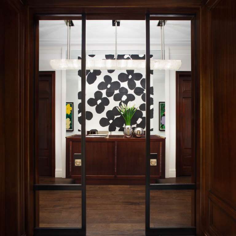 View of a modern office entrance with a wooden reception desk, a black and white abstract wall art, and potted plants. The floor is a combination of dark wood and white marble.