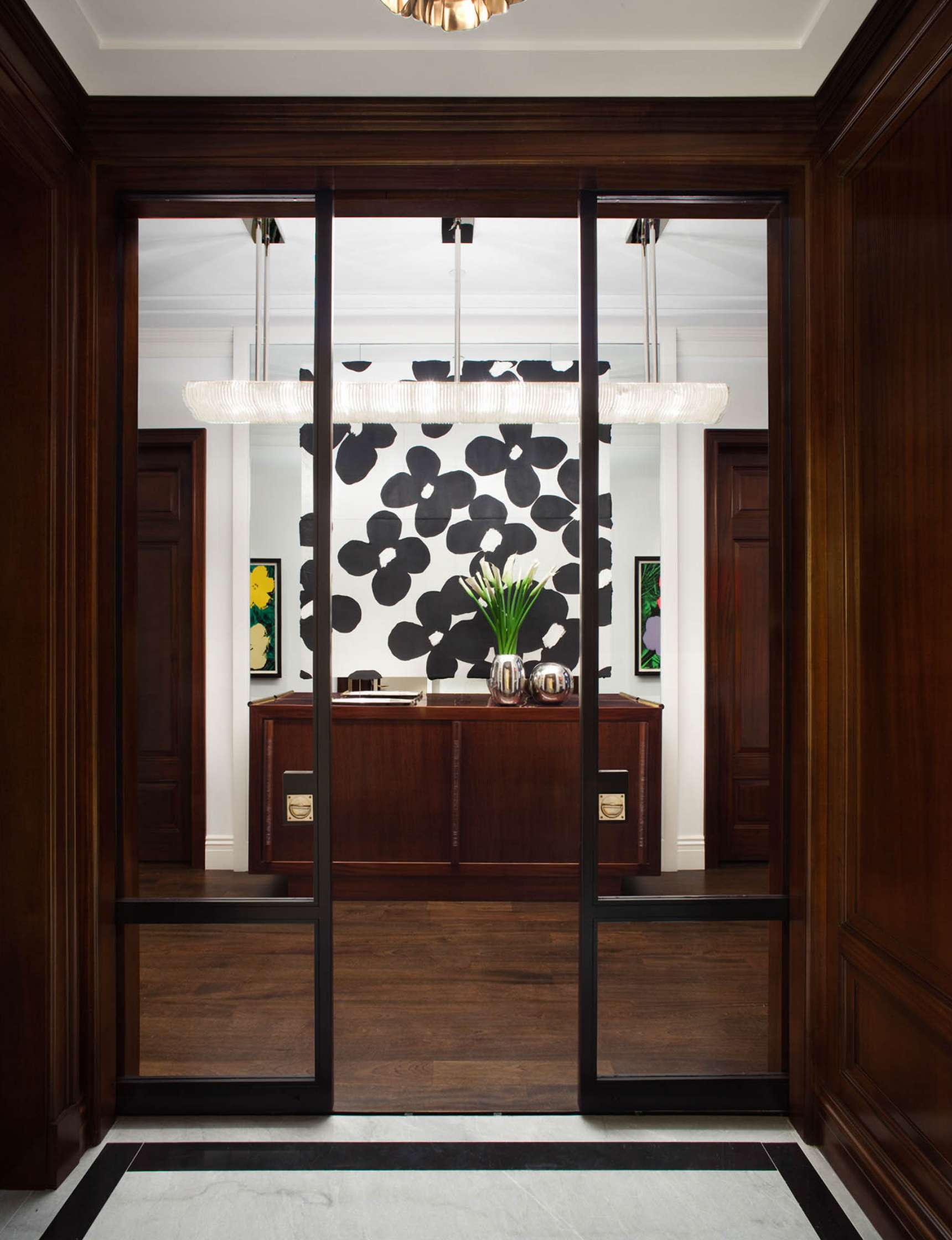 View of a modern office entrance with a wooden reception desk, a black and white abstract wall art, and potted plants. The floor is a combination of dark wood and white marble.