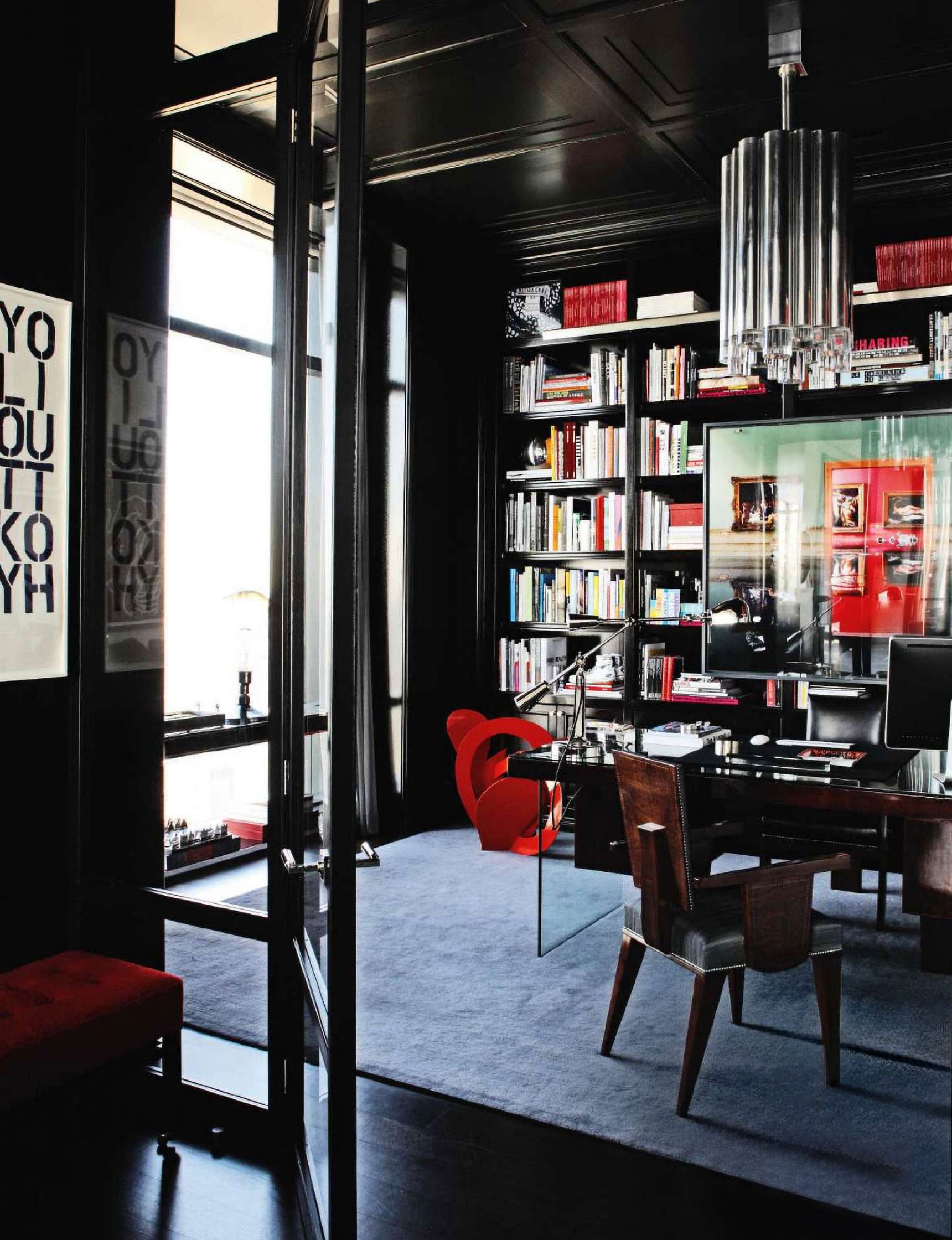 A modern office with a glass desk, wooden chair, and bookshelves filled with books. A red chair and red accents complement the dark walls and ceiling. Large windows allow natural light in.