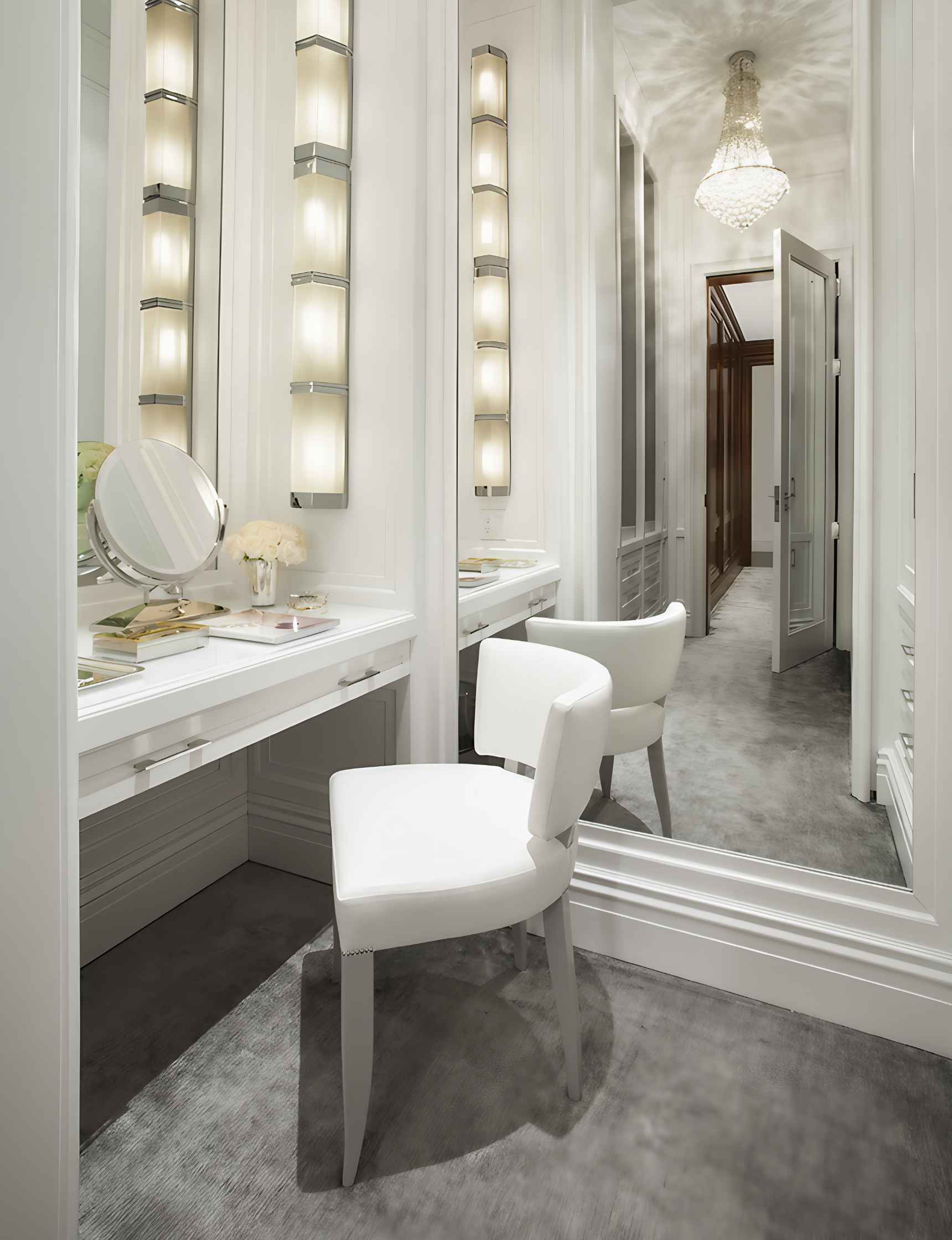 A white makeup vanity with a mirror, chair, and shelves with lights. A hallway with a chandelier is seen in the reflection.