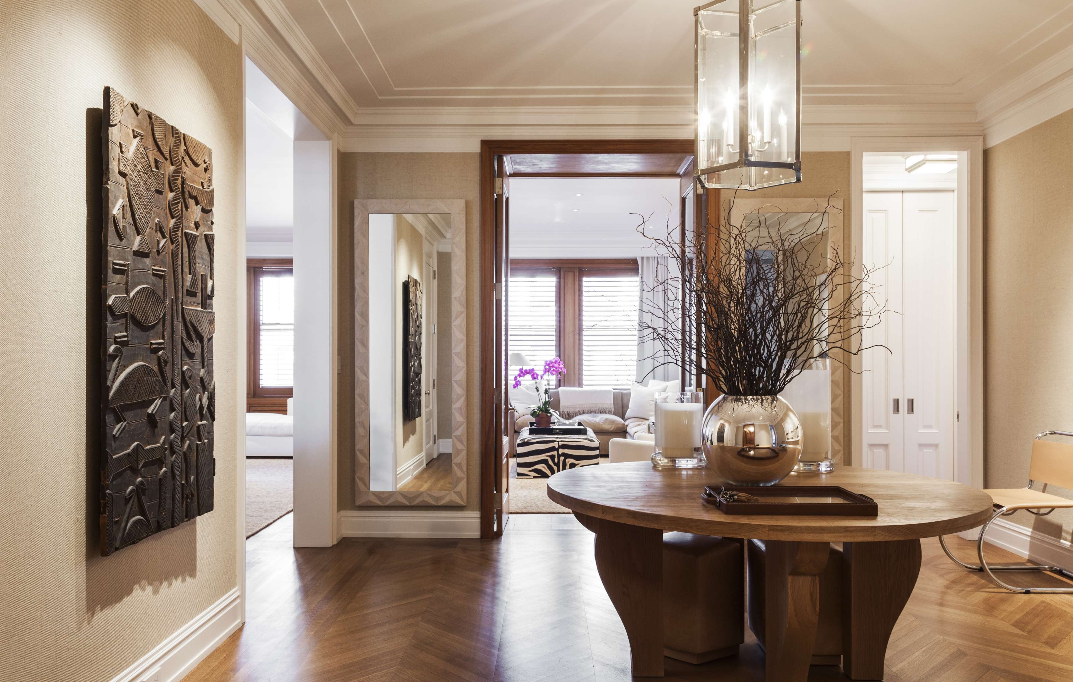 Elegant hallway with a round wooden table, a large mirror, a textured wall art, and a view into a bright living room with a white sofa and striped rug.