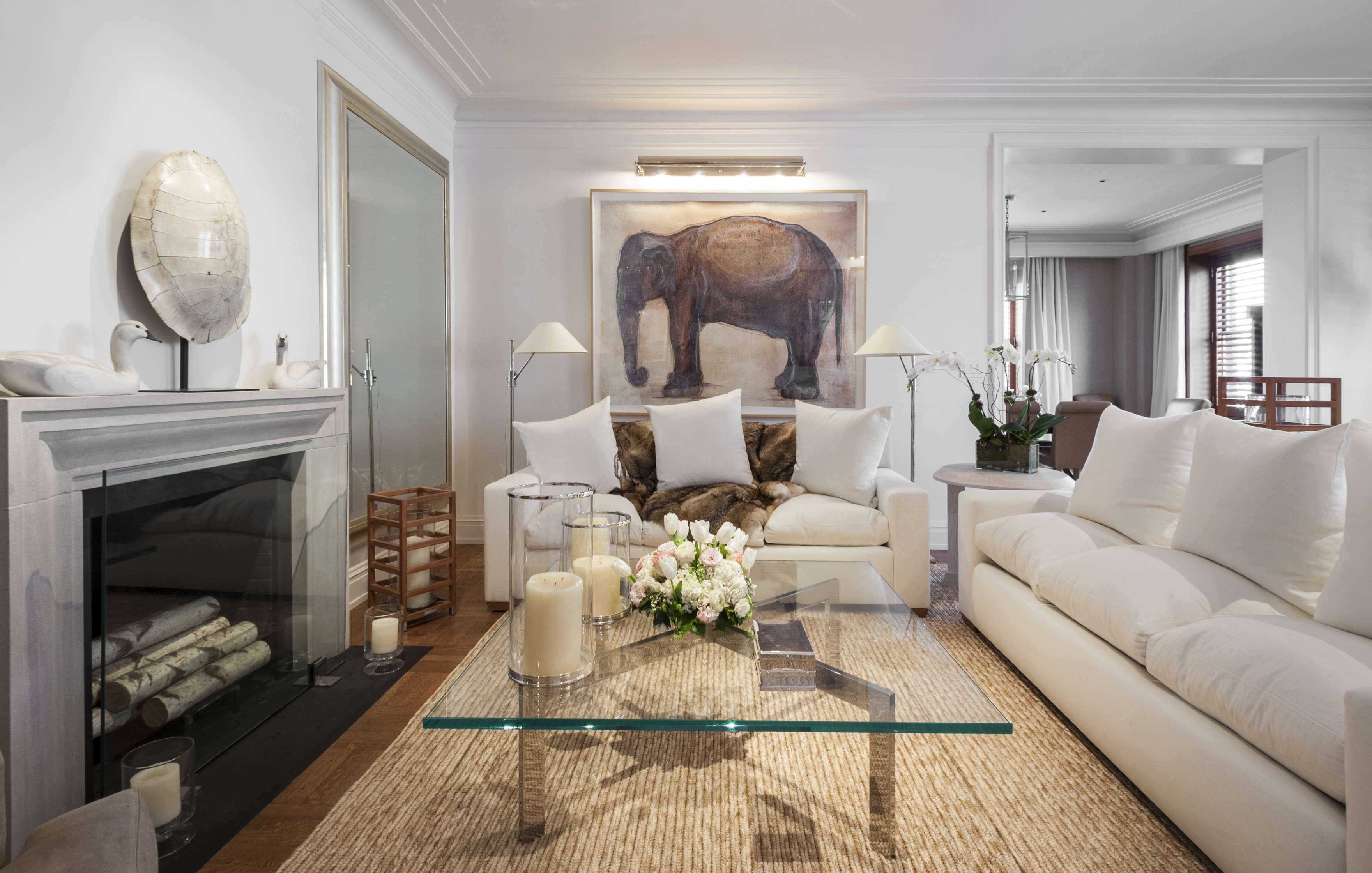 Elegant living room with white sofas, a glass coffee table, and a large elephant painting. The room has a fireplace, decorative candles, and soft lighting. Flowers are arranged on the table.