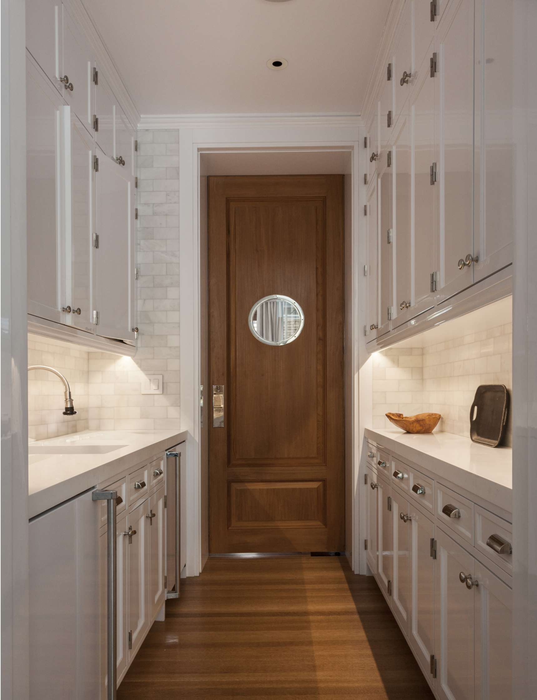 A narrow galley kitchen with white cabinets, wooden flooring, under-cabinet lighting, and a wooden door featuring a small round window.