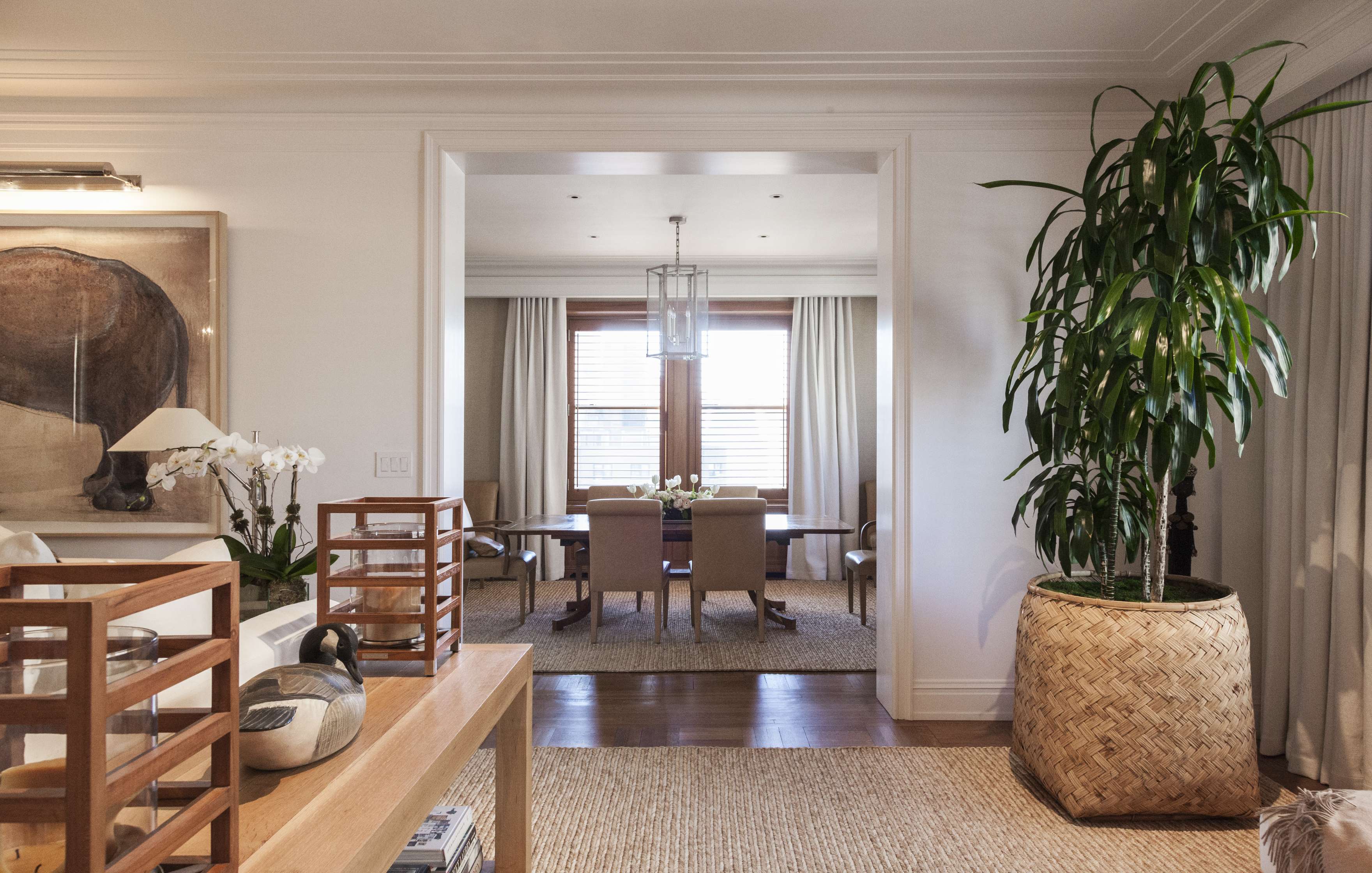 A living room with wood accents opens to a dining area. A large plant is on the right, and a wooden table with chairs is visible in the background.