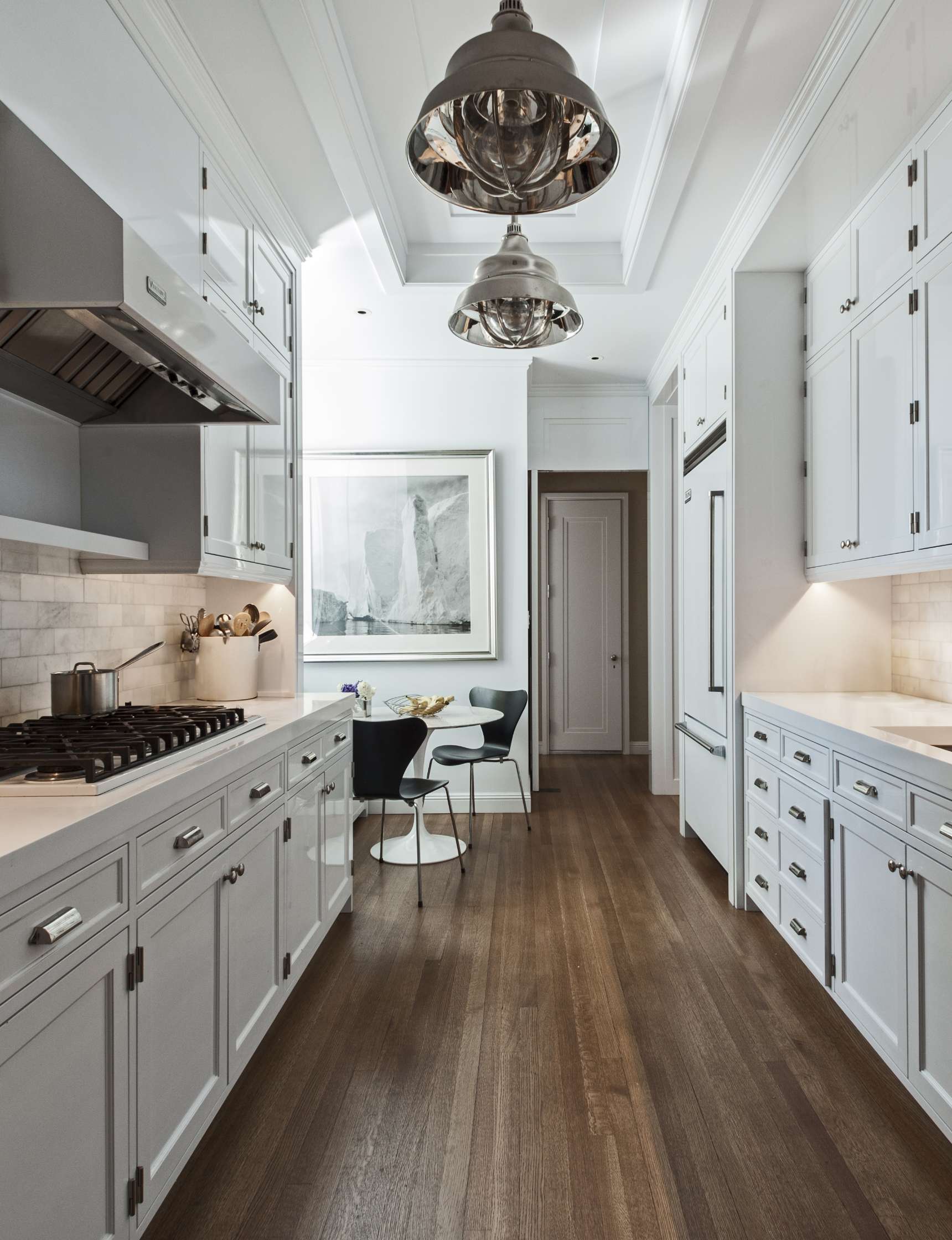 Modern kitchen with white cabinets, stainless steel appliances, and wooden flooring. A small dining area with a round table and two chairs is visible next to a large framed artwork.