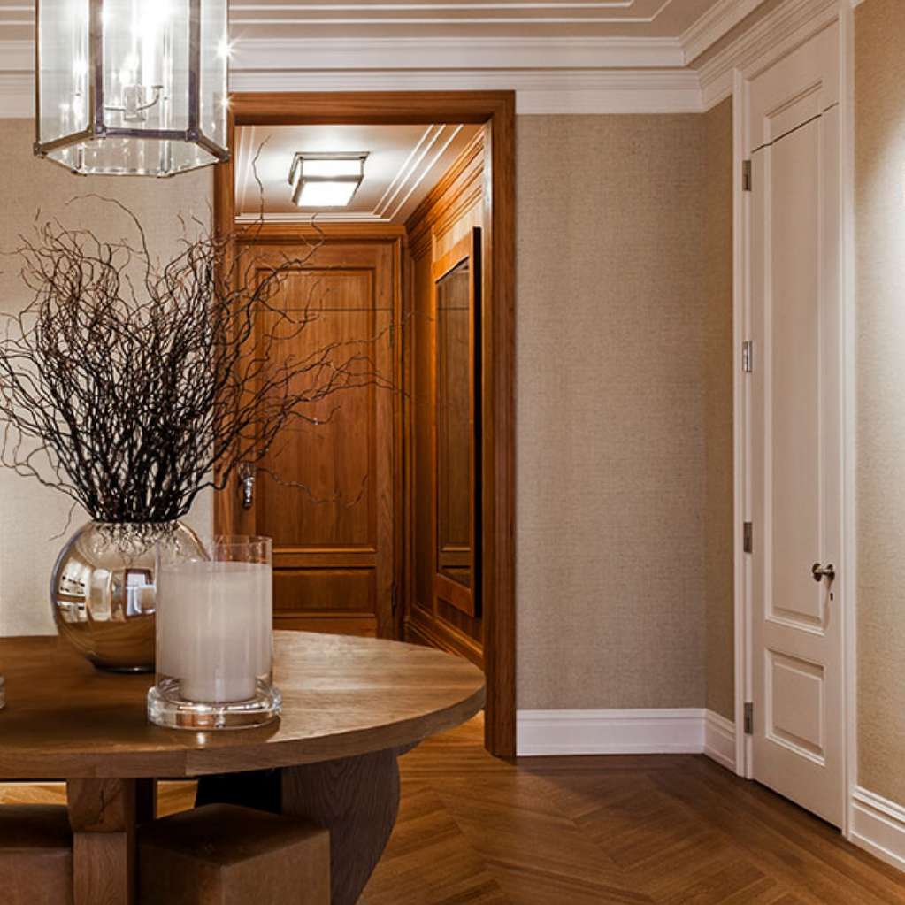 Elegant hallway with a round wooden table, decorative branches in a vase, two large candles, modern artwork on walls, leather chair, and warm lighting.