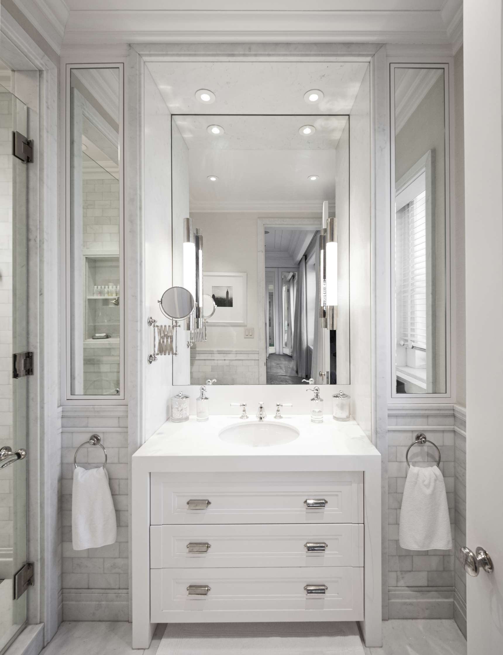 A small, elegant bathroom with a white vanity, three drawers, a large mirror, wall-mounted lights, and a glass shower door. Towels hang on both sides of the vanity.