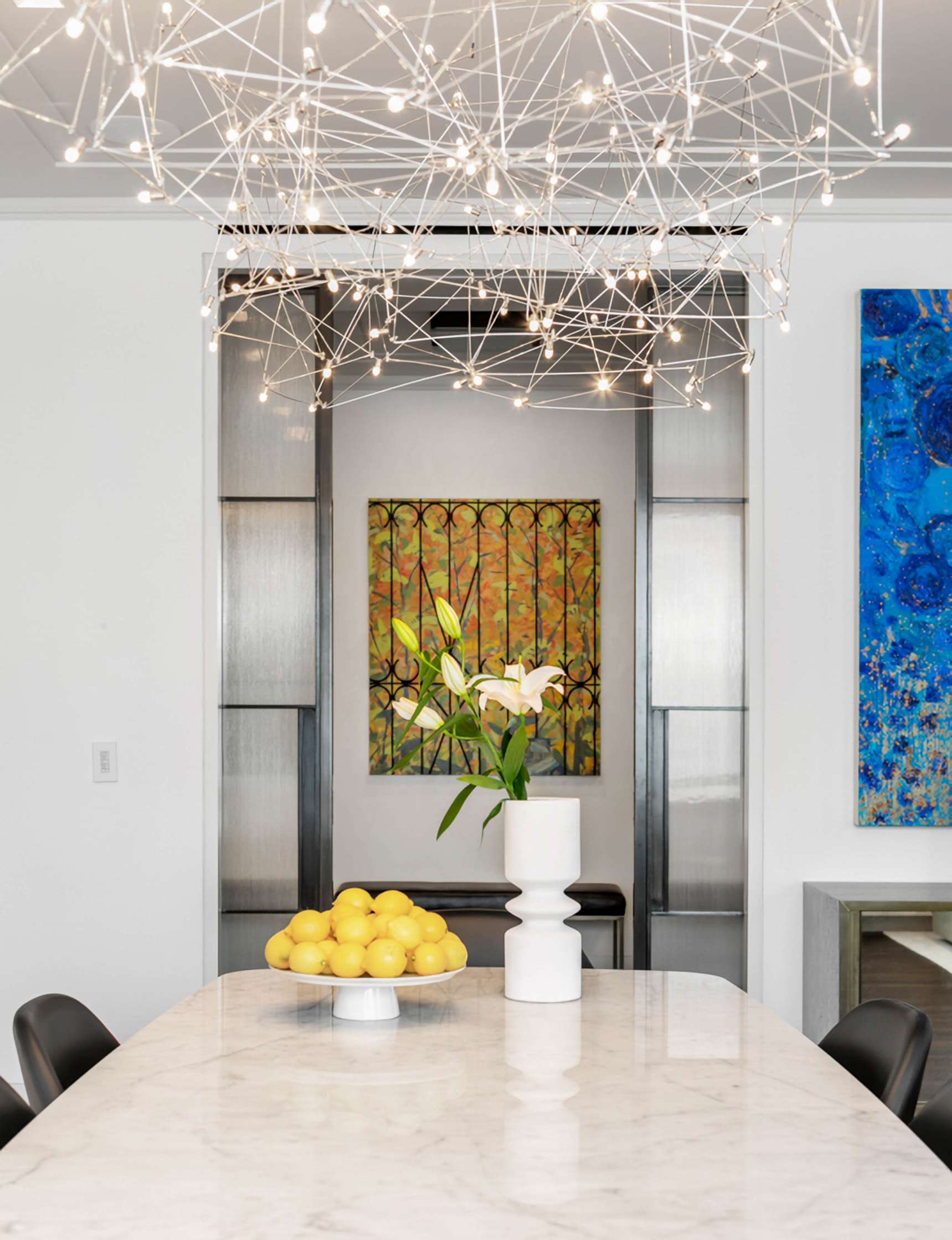 A modern dining room with a marble table, a bowl of lemons, a white vase with white lilies, two paintings, and a decorative light fixture hanging above.