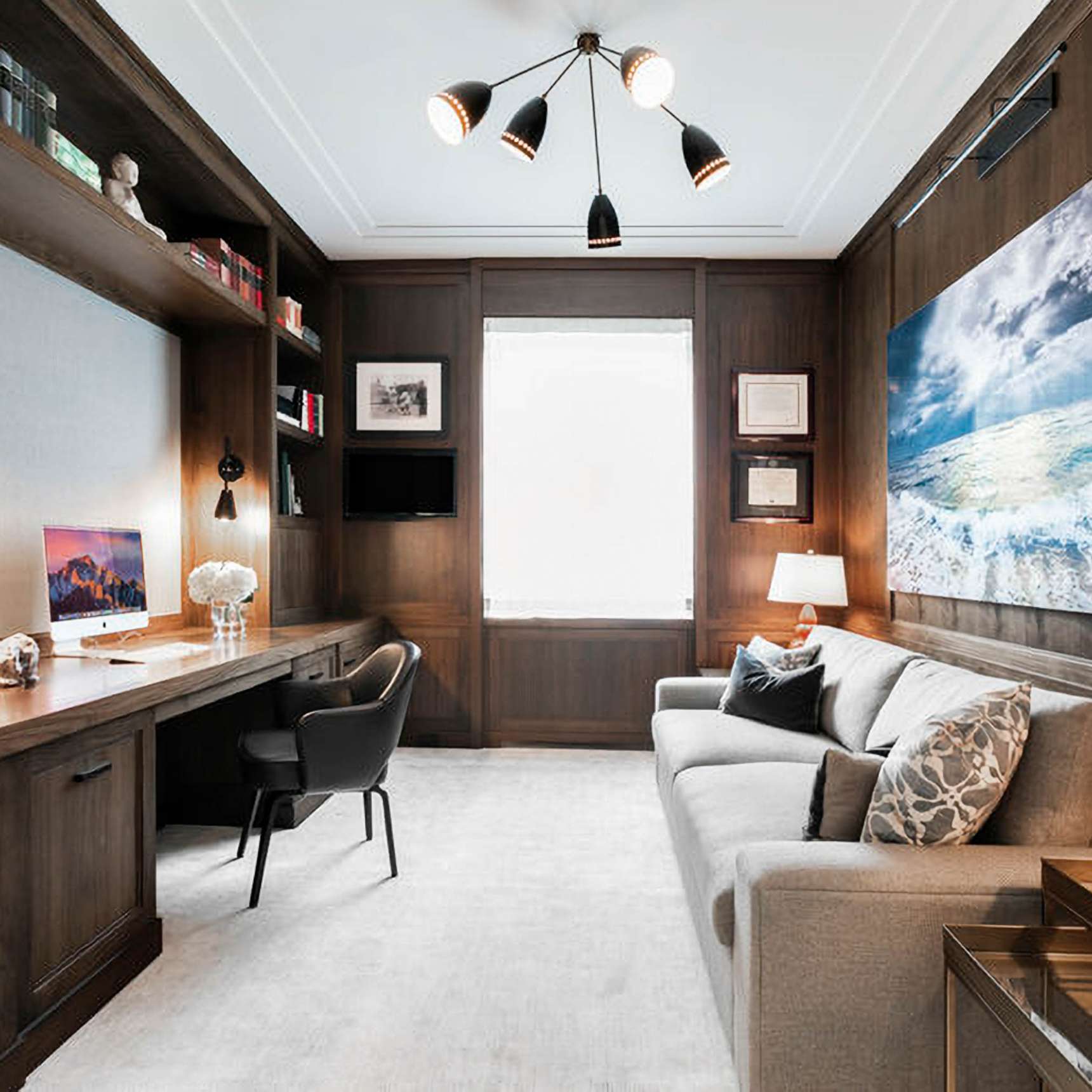 A modern home office with wooden shelves, a black chair at a desk with a computer and lamp, a gray sofa, a large seascape artwork, and a window in the center. A modern ceiling light fixture hangs above.